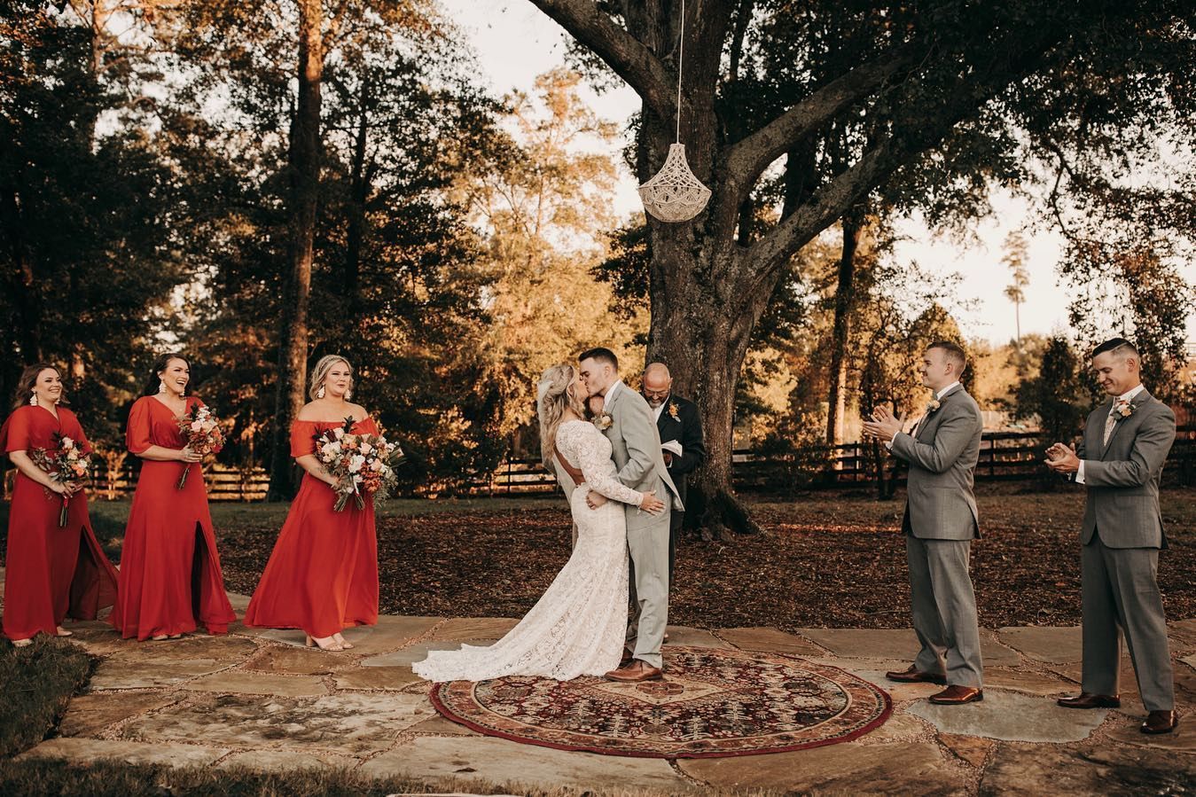 Wedding ceremony: couple kissing, bridesmaids in red, groomsmen in gray suits, outdoors near a tree.