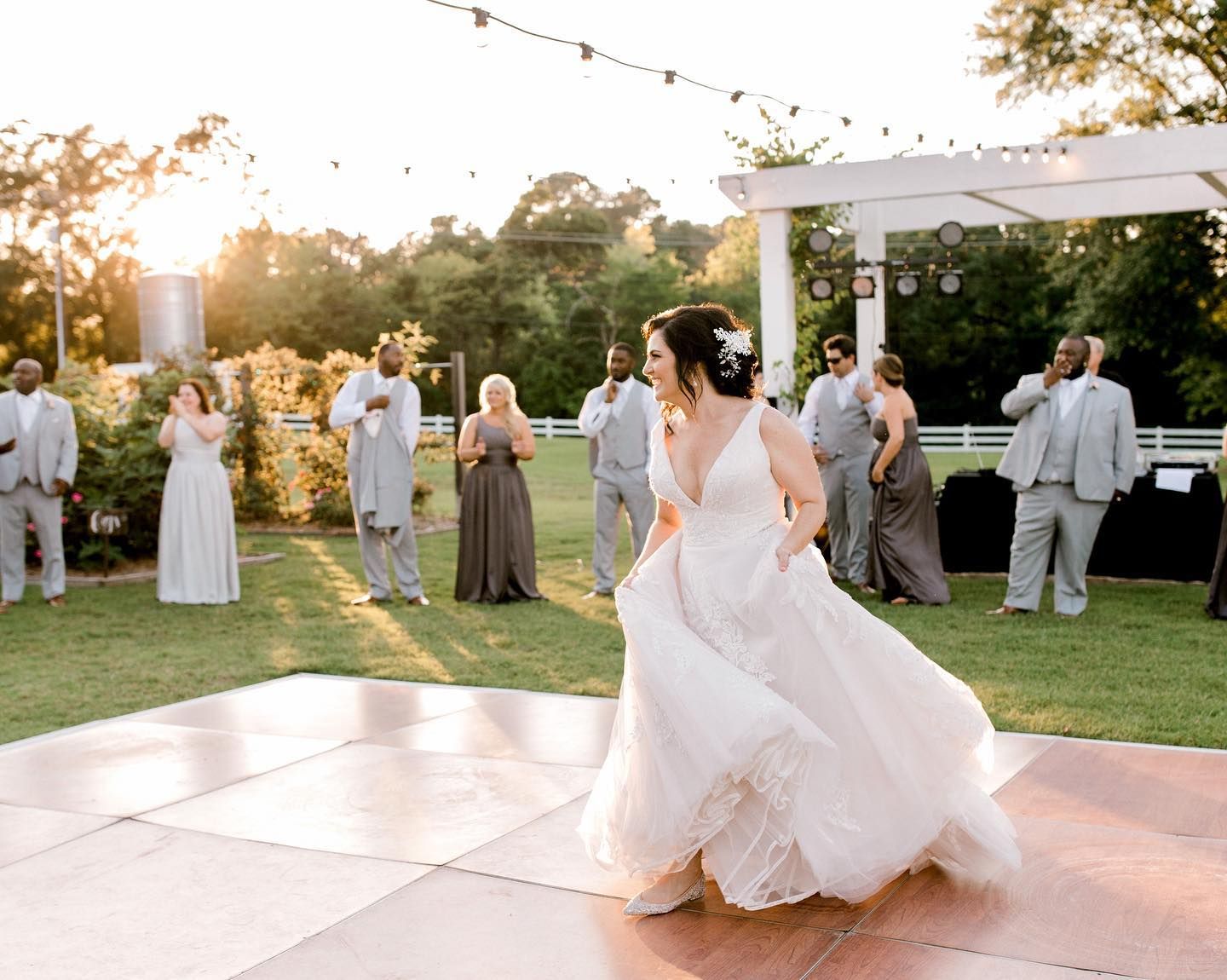 Bride dancing on a checkered dance floor at an outdoor wedding. Sun sets in background. Wedding party watches.
