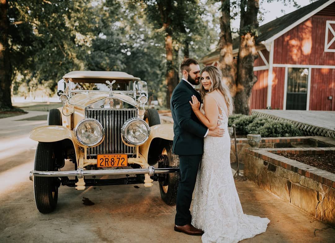 Bride and groom embrace by a yellow vintage car at their wedding. Red barn in the background.
