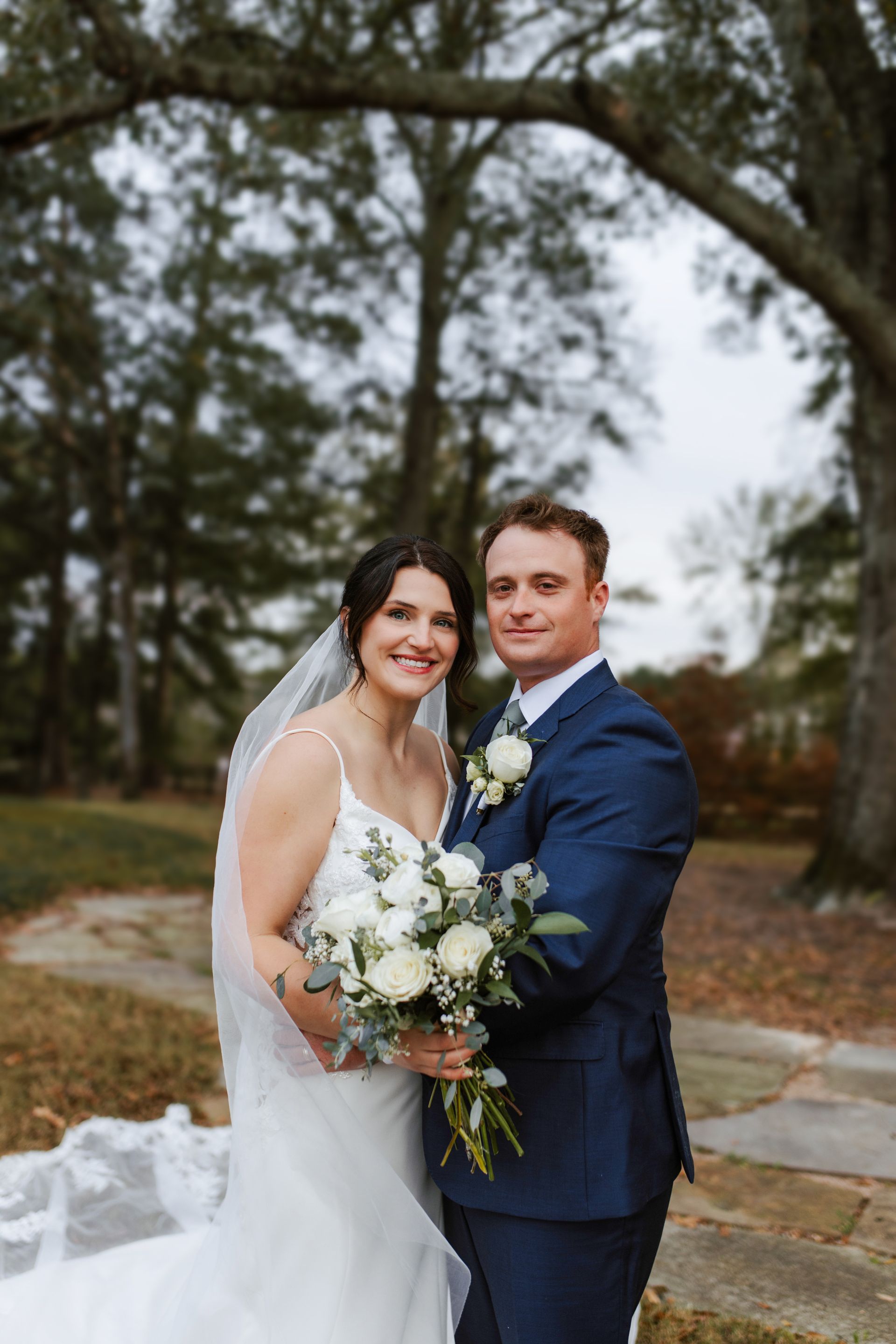 Wedding couple smiling outdoors; the bride holds a bouquet, the groom wears a blue suit, trees in the background.