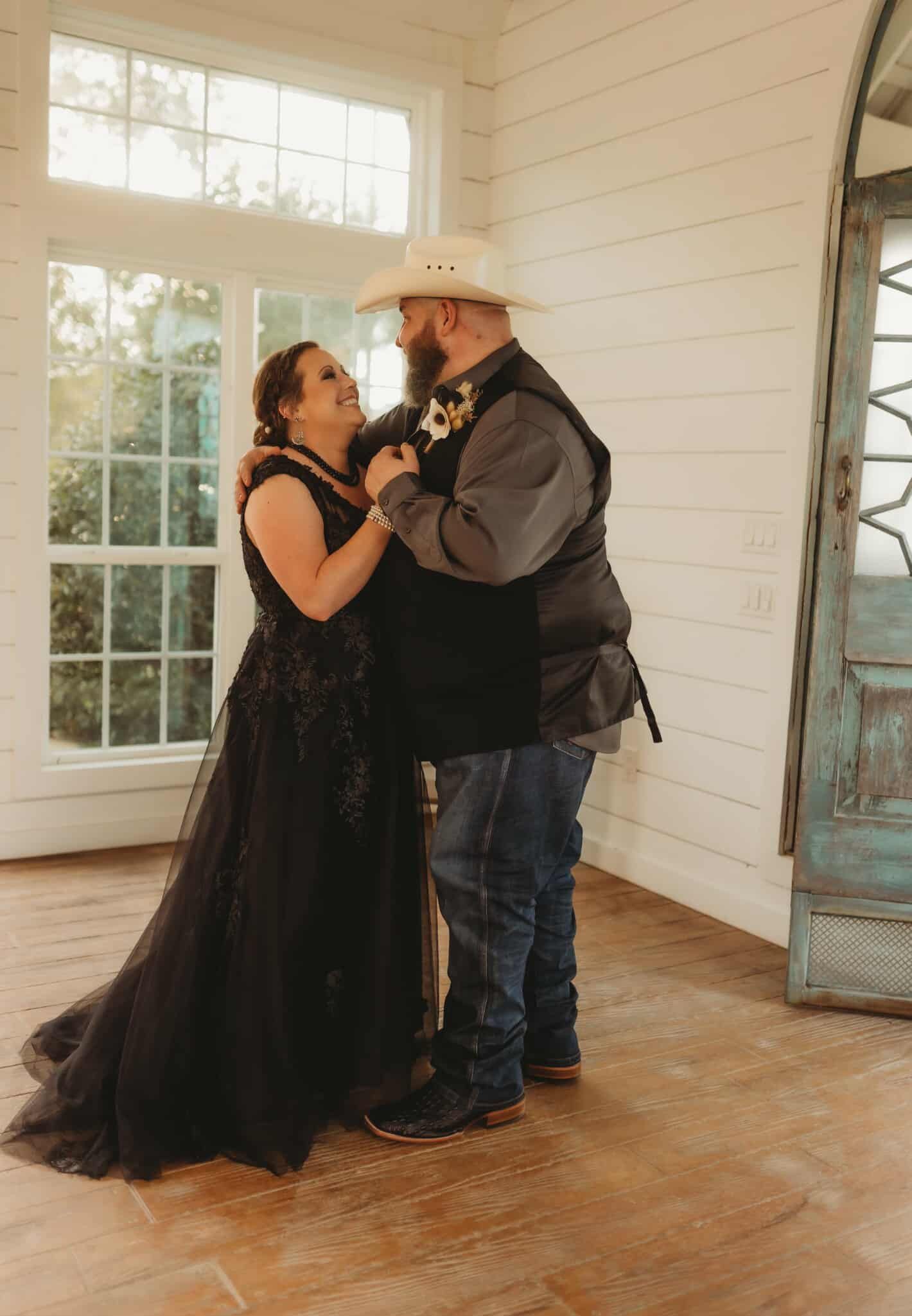 Couple dancing, bride in black gown, groom in vest and cowboy hat, in a sunlit room.