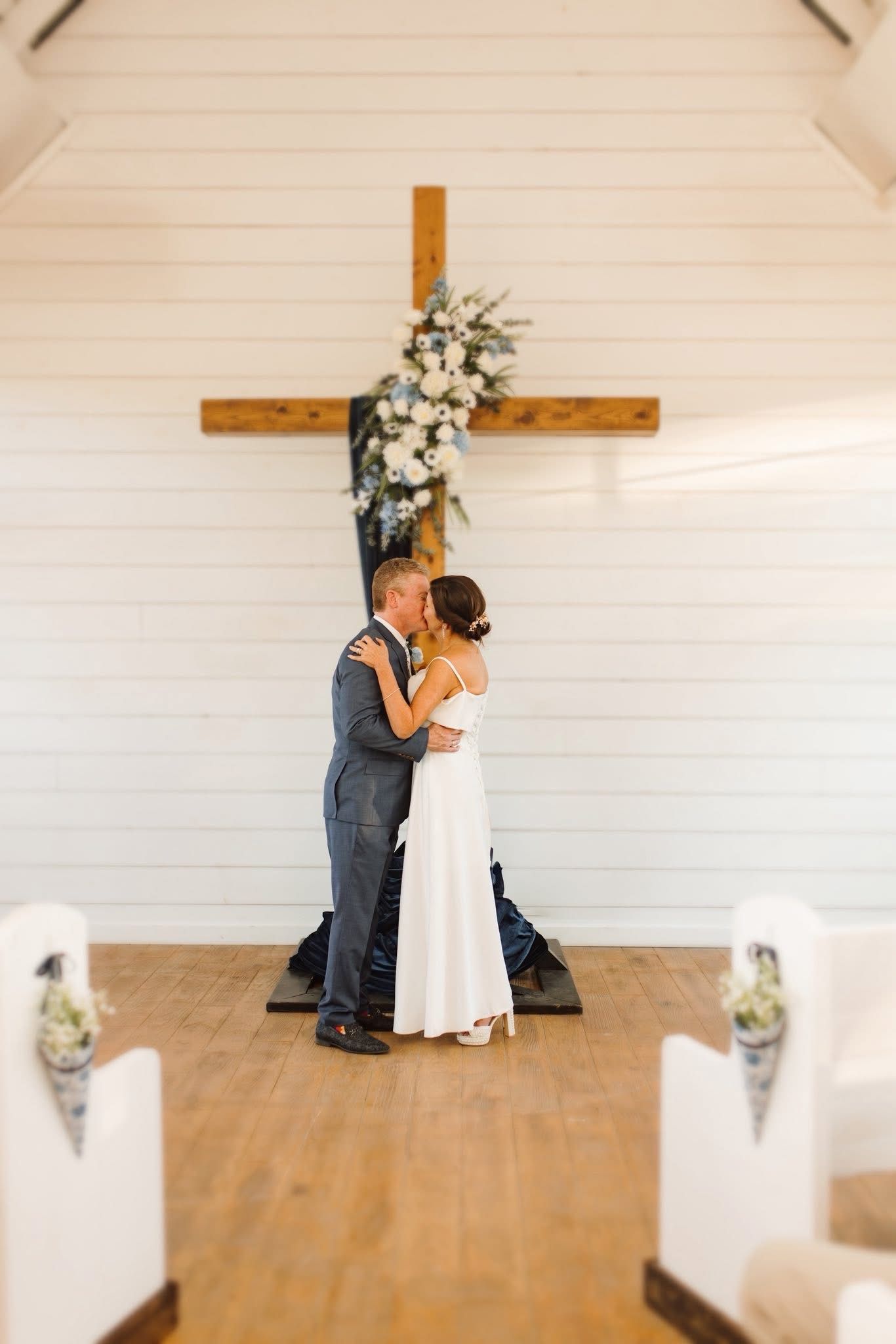 Couple kissing in front of wooden cross with floral arrangement; white building interior.