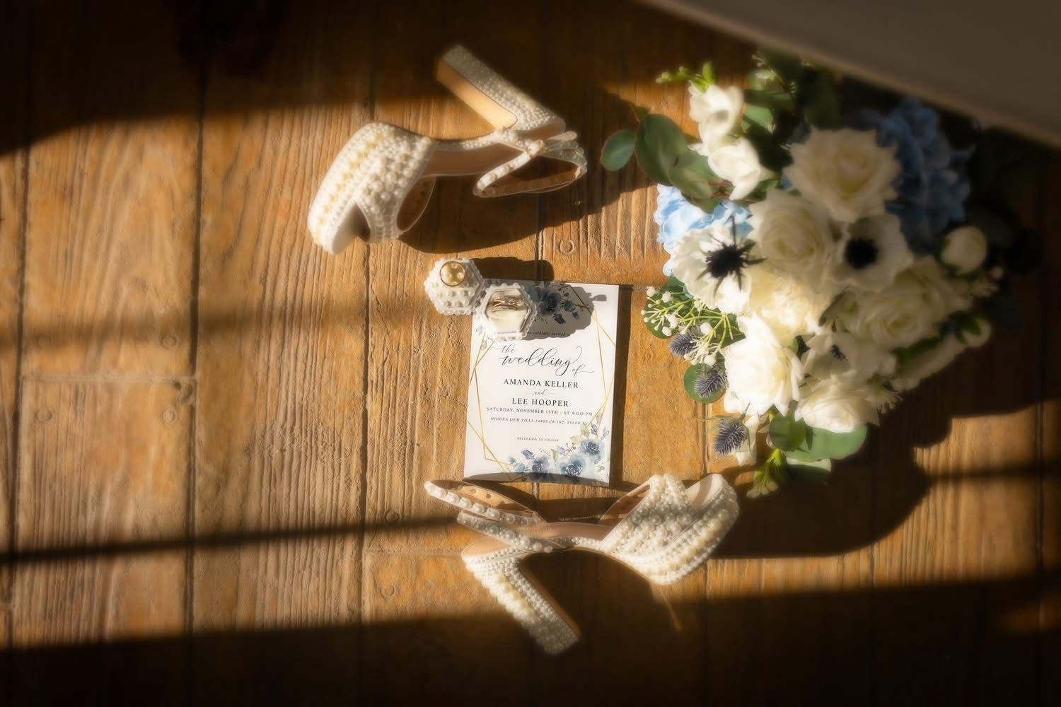 Wedding details: shoes, invitation, bouquet on wooden floor, lit by sunlight.