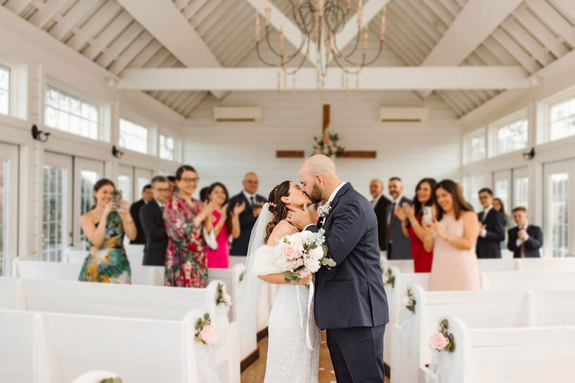 Newlyweds kiss at the altar, guests clap, white chapel with cross and floral decor.