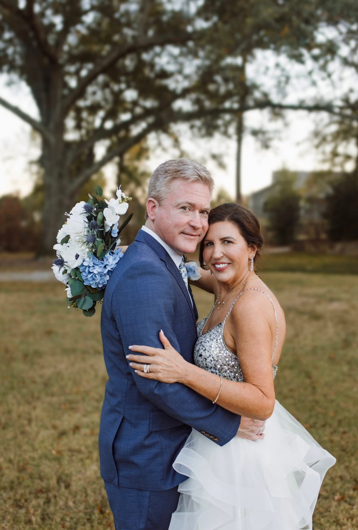 Man in blue suit and woman in white dress embrace outdoors, smiling.