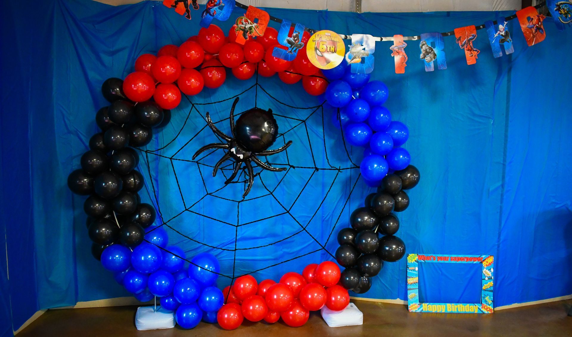 Spider-Man-themed party backdrop. Circular balloon arch with a spider web and banner. Red, blue, black balloons.