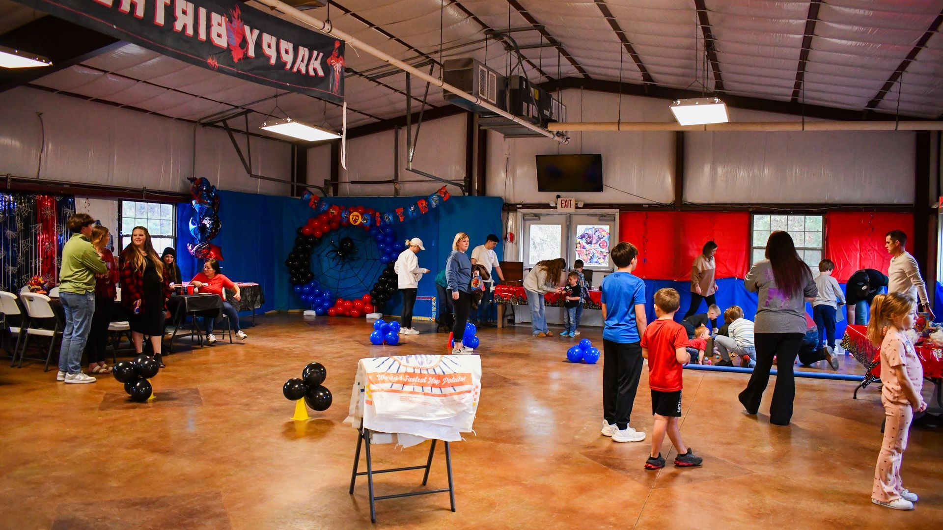 Birthday party inside a large open space, decorated with balloons and streamers. People mingle, some children play.