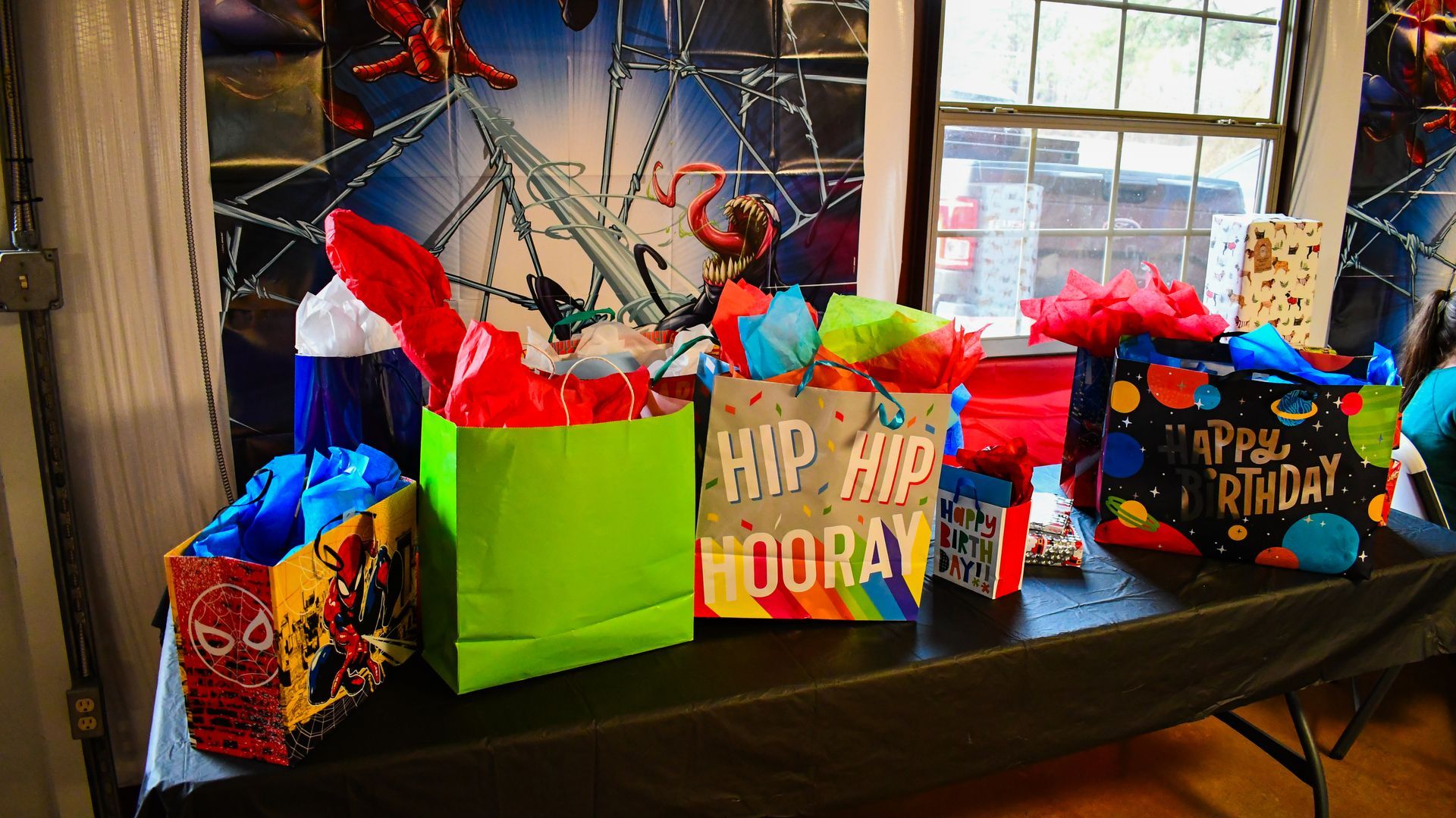 Birthday gifts on a table, with a Spider-Man backdrop. Colorful gift bags with tissue paper and decorations.