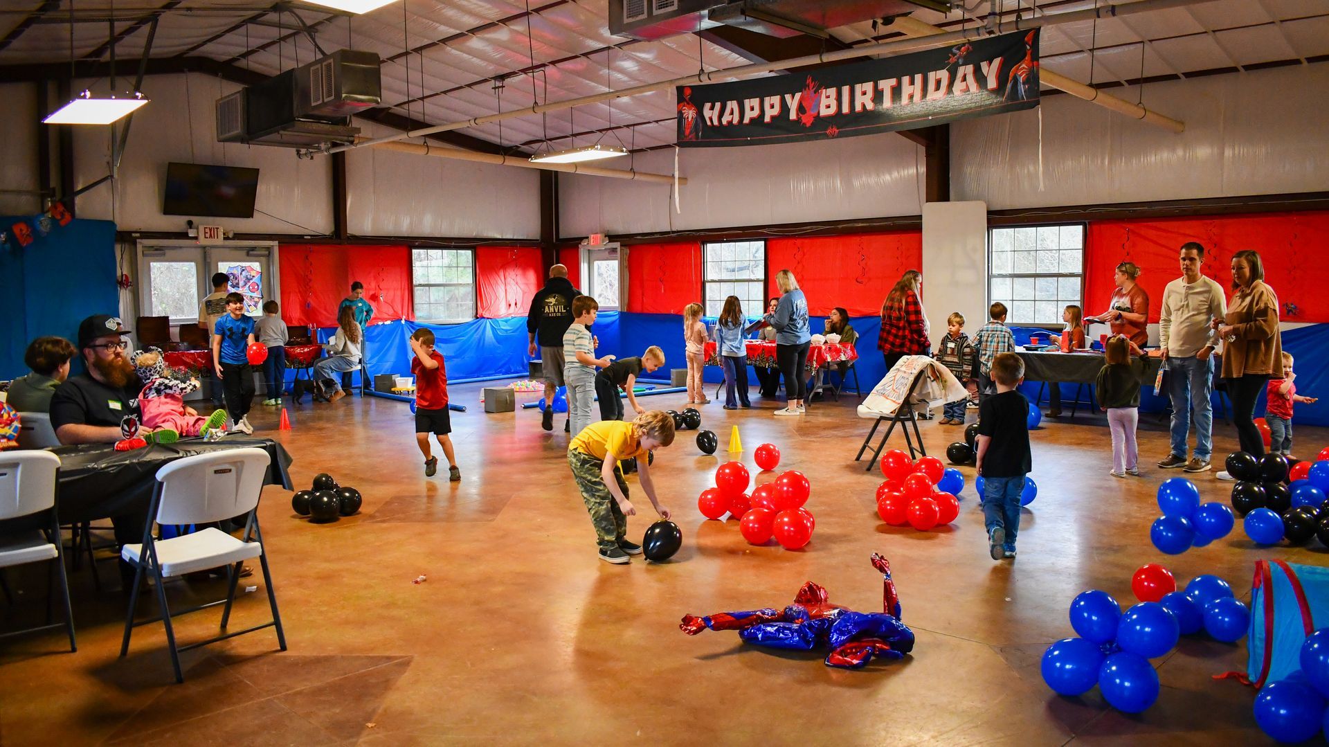 Birthday party in a large hall with balloons, tables, and people celebrating. Banner reads 