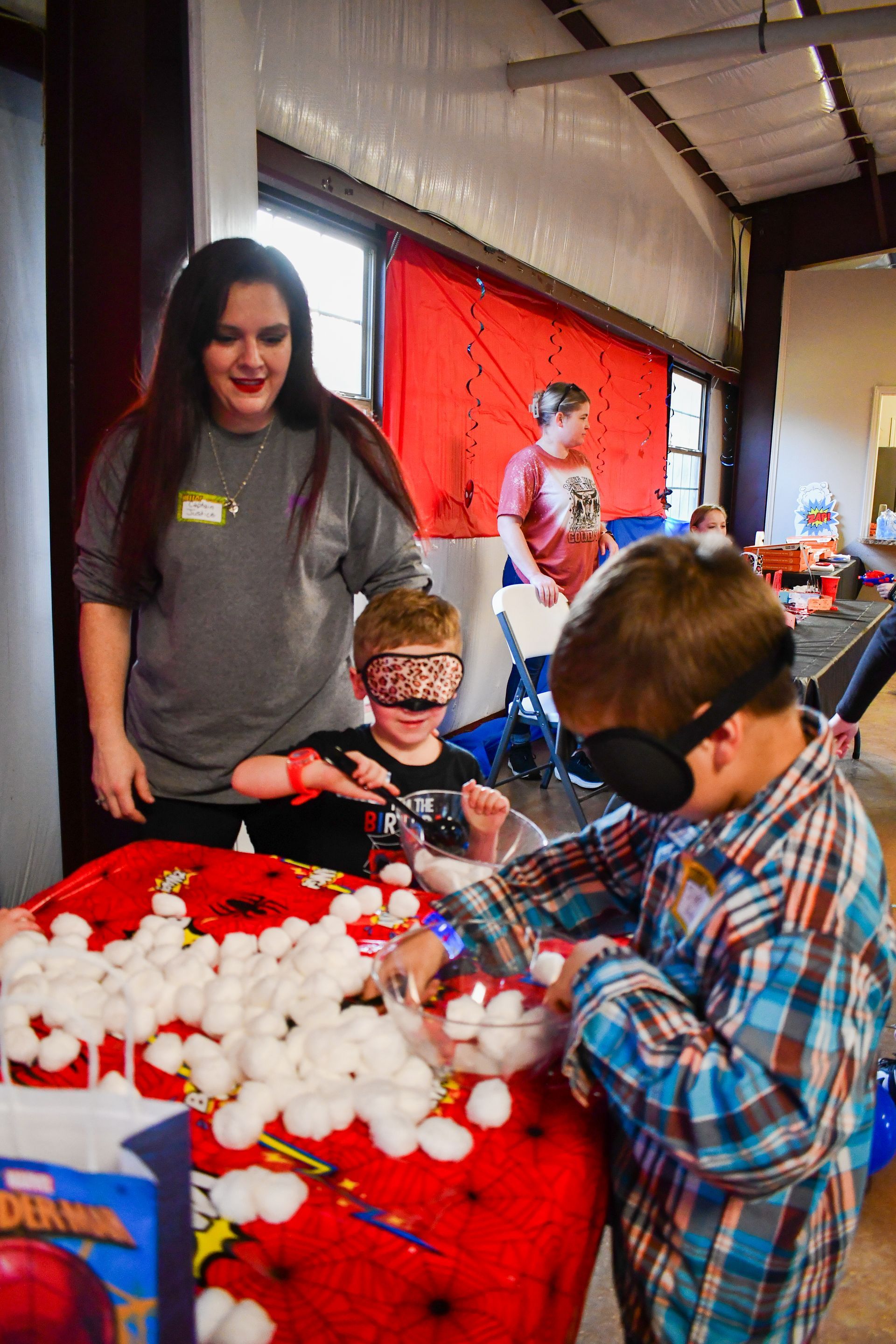 Two blindfolded children at a table, touching cotton balls while an adult watches. Red table cloth.