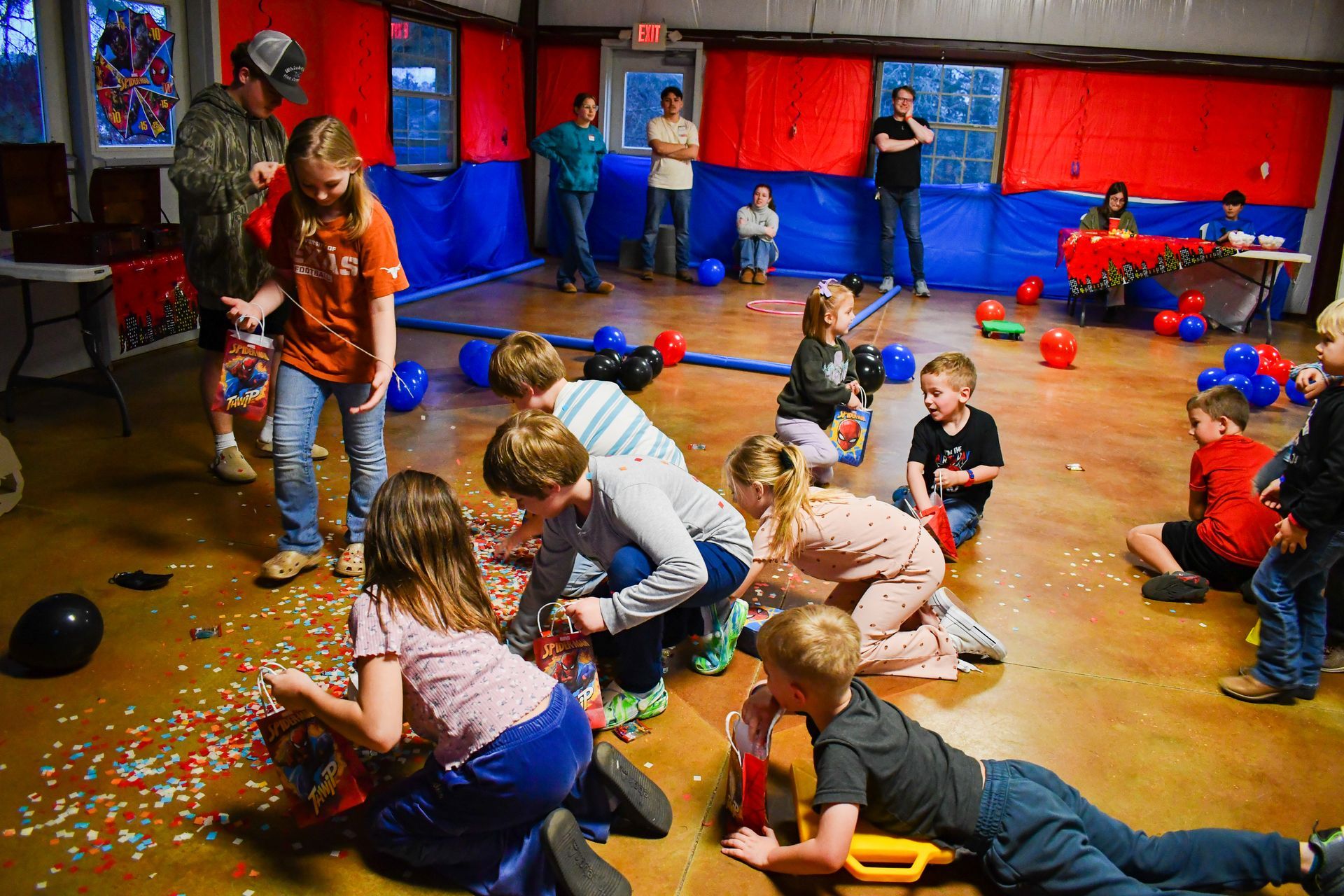 Children open party favors on a floor, with scattered confetti and balloons; adults watch nearby in a decorated room.