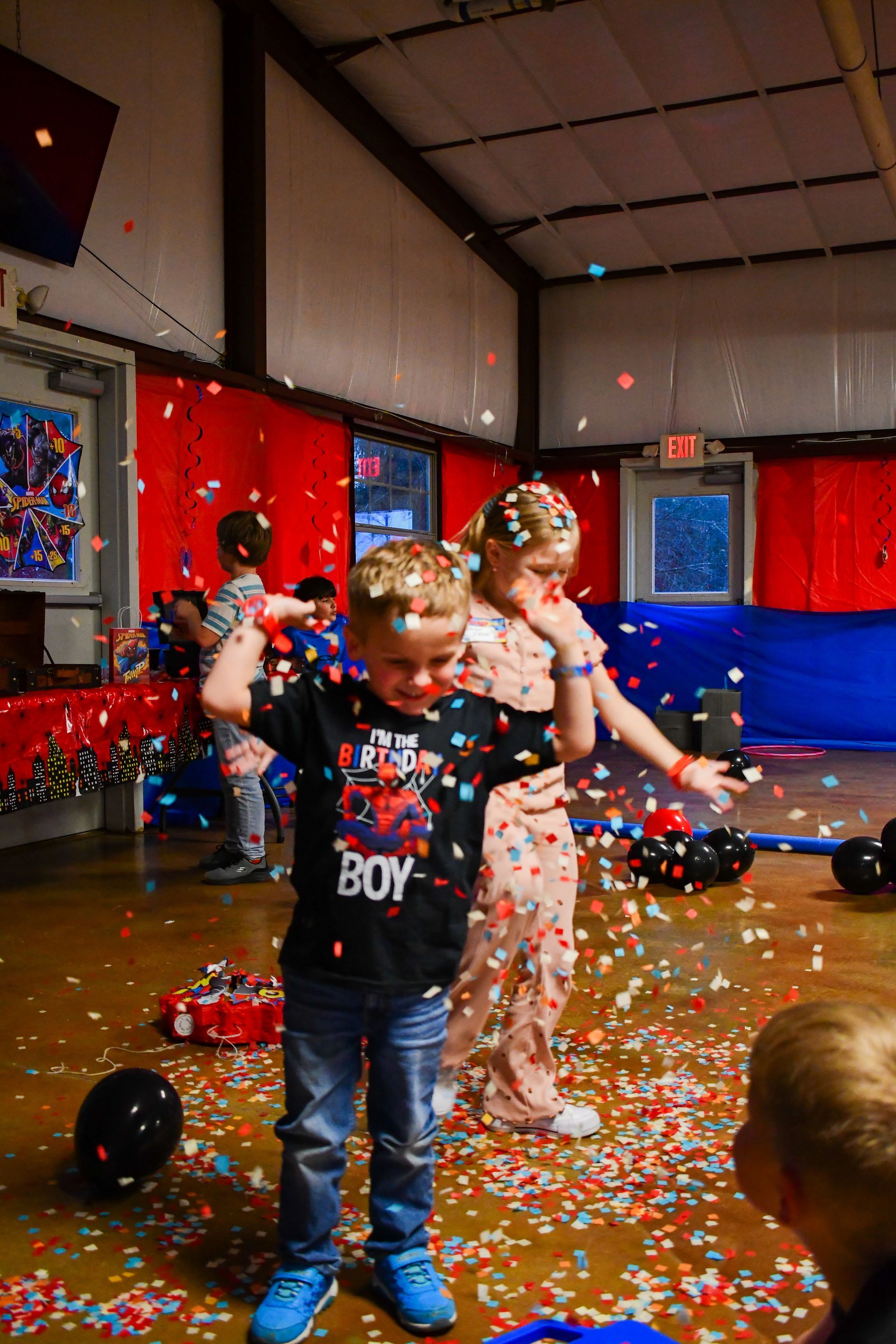 Boy in confetti-filled room, celebrating. Red, blue decor. Smiling, arms raised. Another person with them.