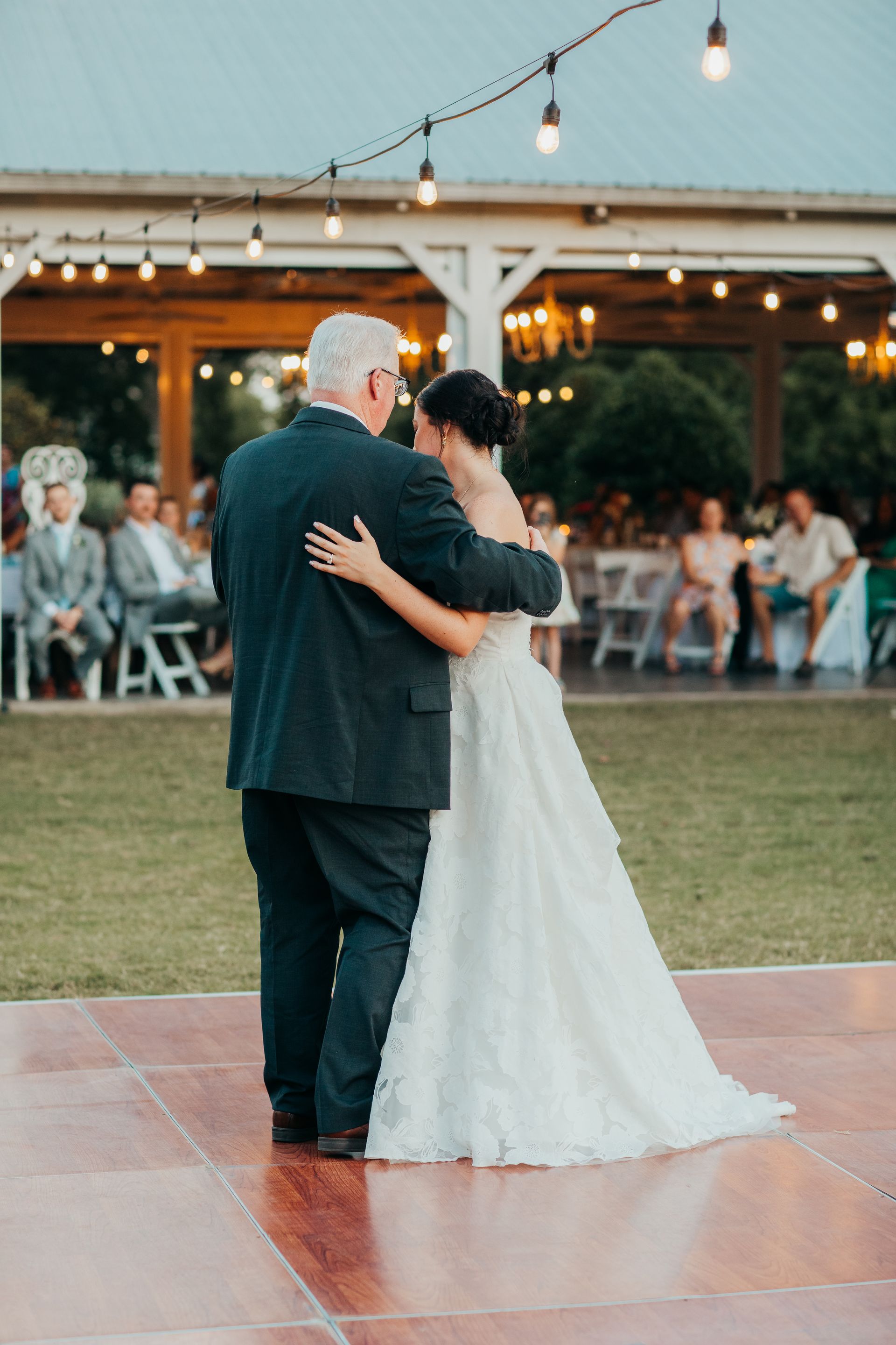 Father and bride dance outdoors, under string lights. Bride wears white gown, man wears suit.