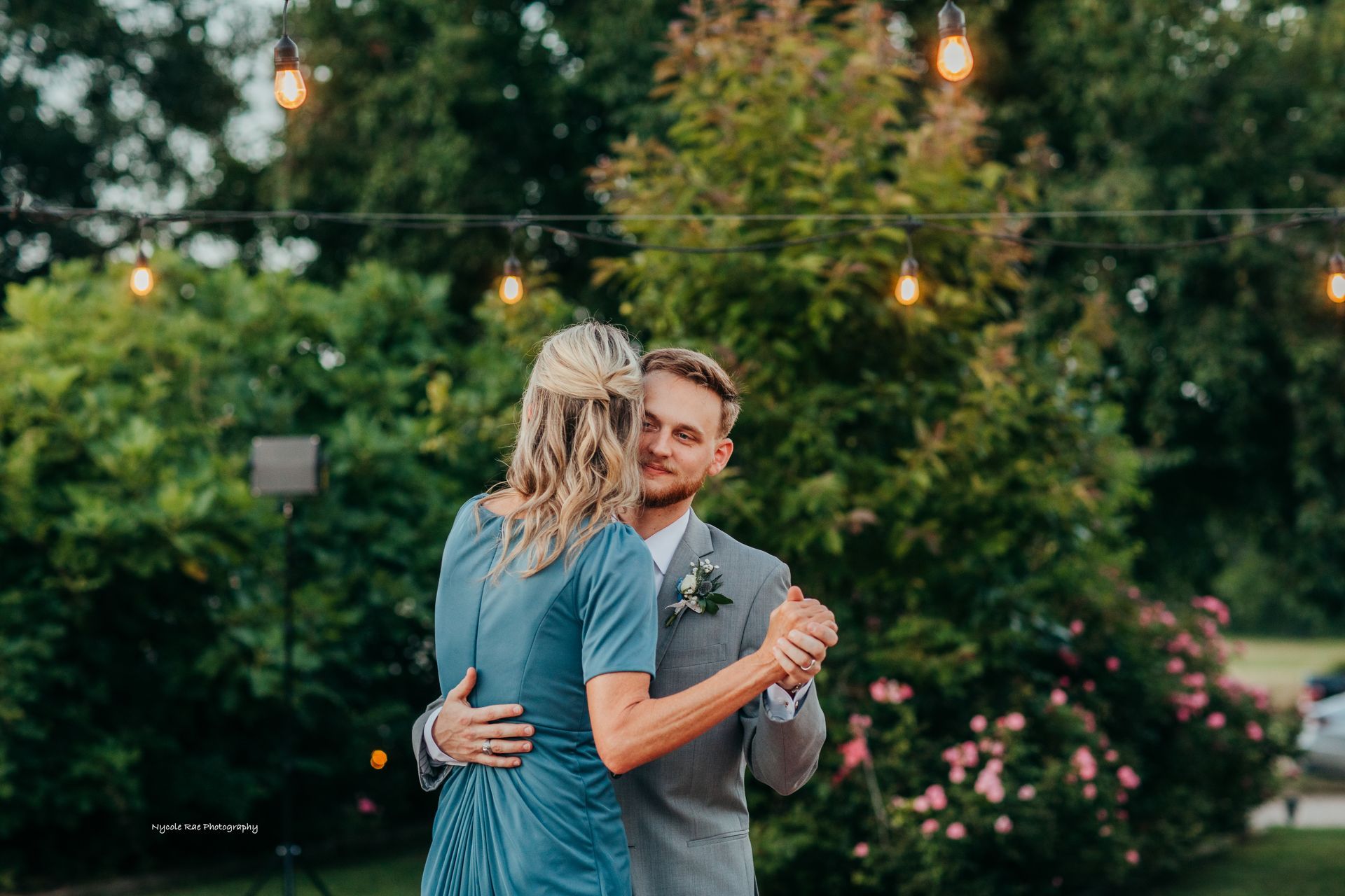 Man dancing with a woman outside at a celebration, string lights overhead, greenery in the background.