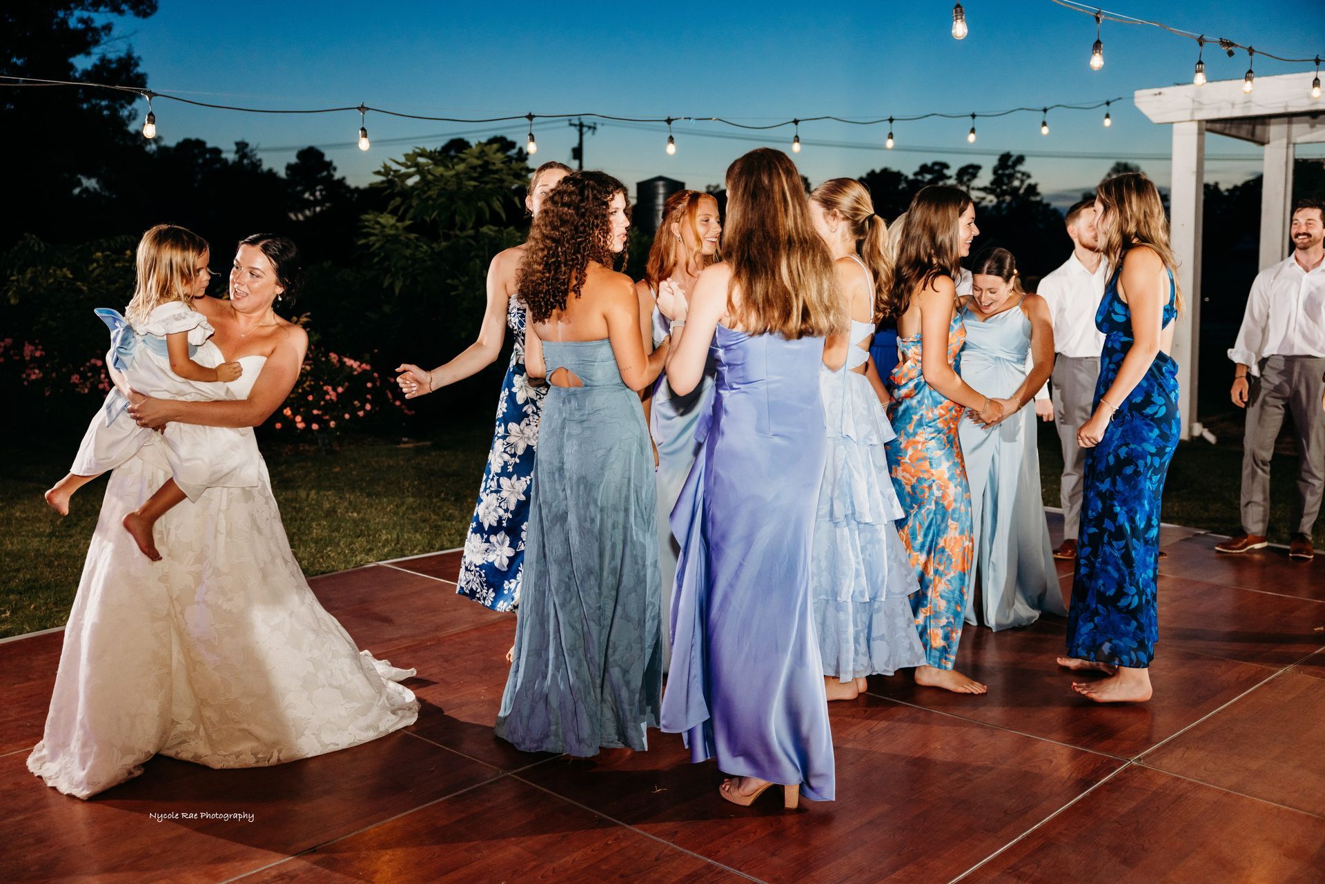 Bride holds child, surrounded by bridesmaids in blue dresses on a dance floor at dusk.