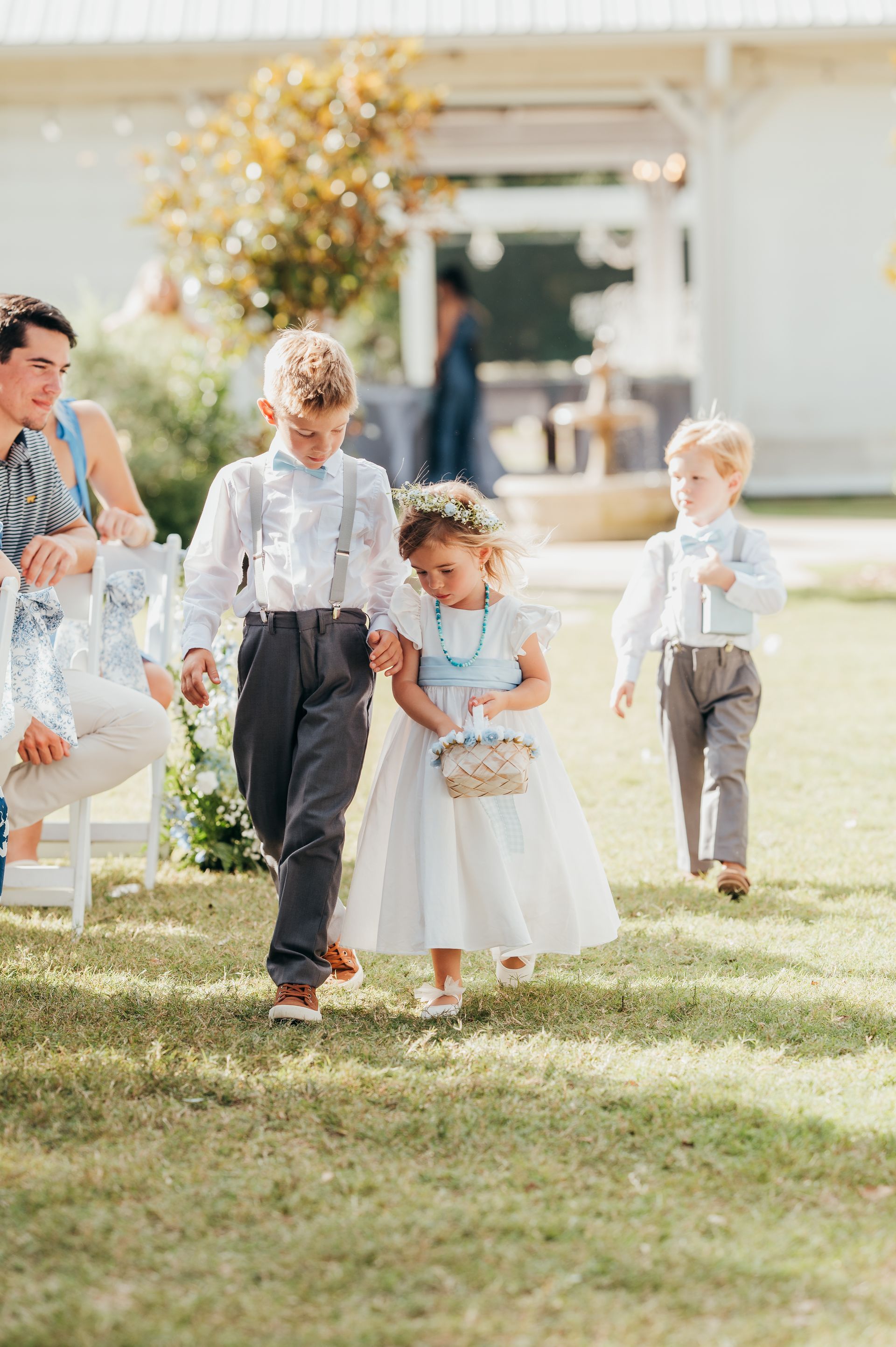 Children walking down a grassy aisle. A boy holds the hand of a girl carrying flowers, another boy follows.