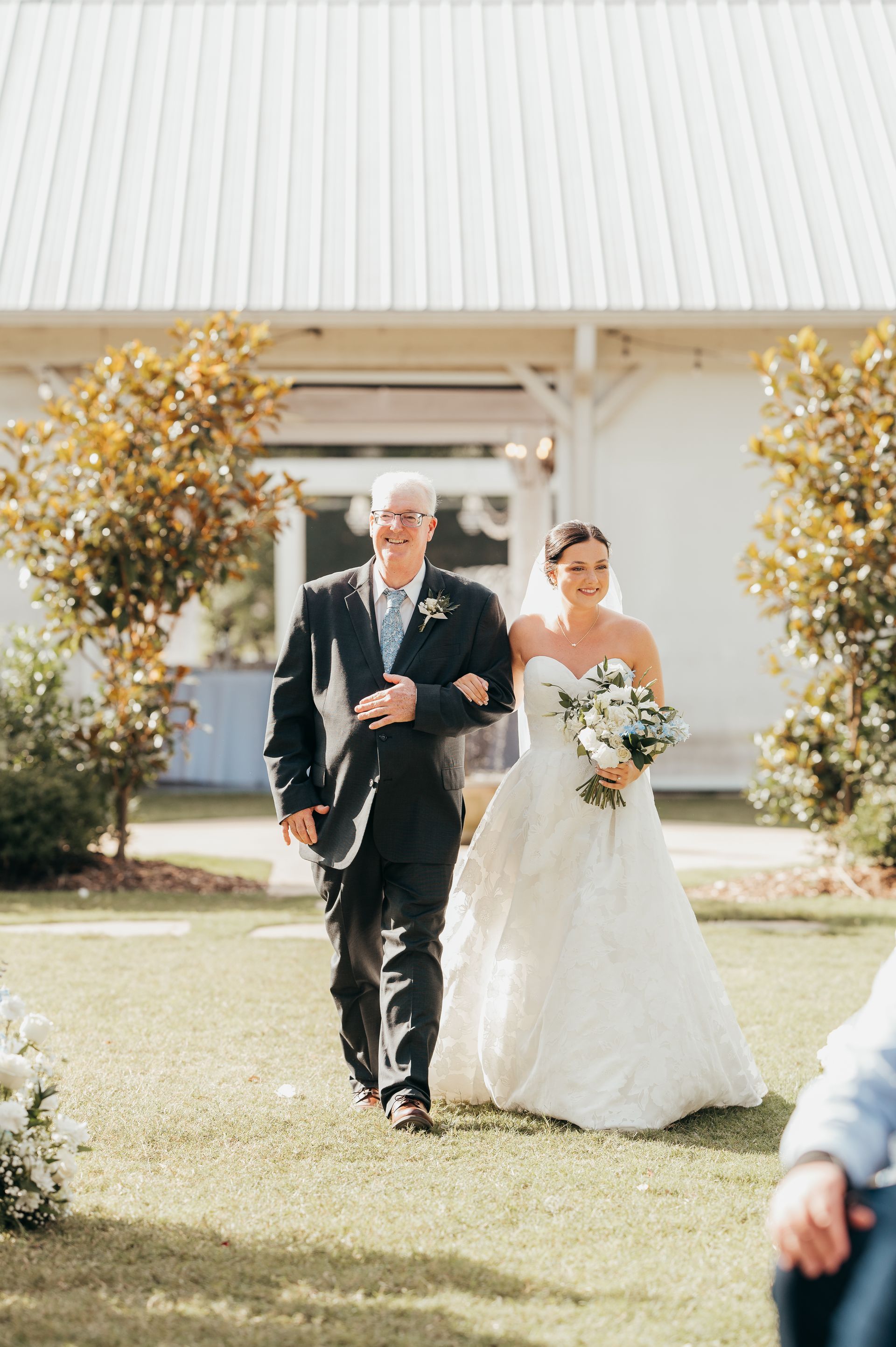 Bride and father walk down aisle at outdoor wedding. Bride holds bouquet, wears white dress.