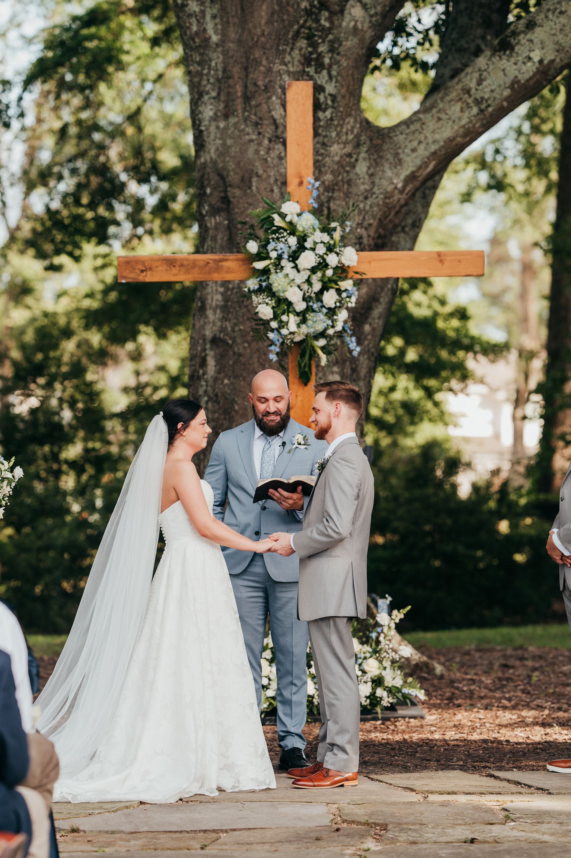 Wedding ceremony: couple holding hands, standing before a wooden cross adorned with flowers outdoors.