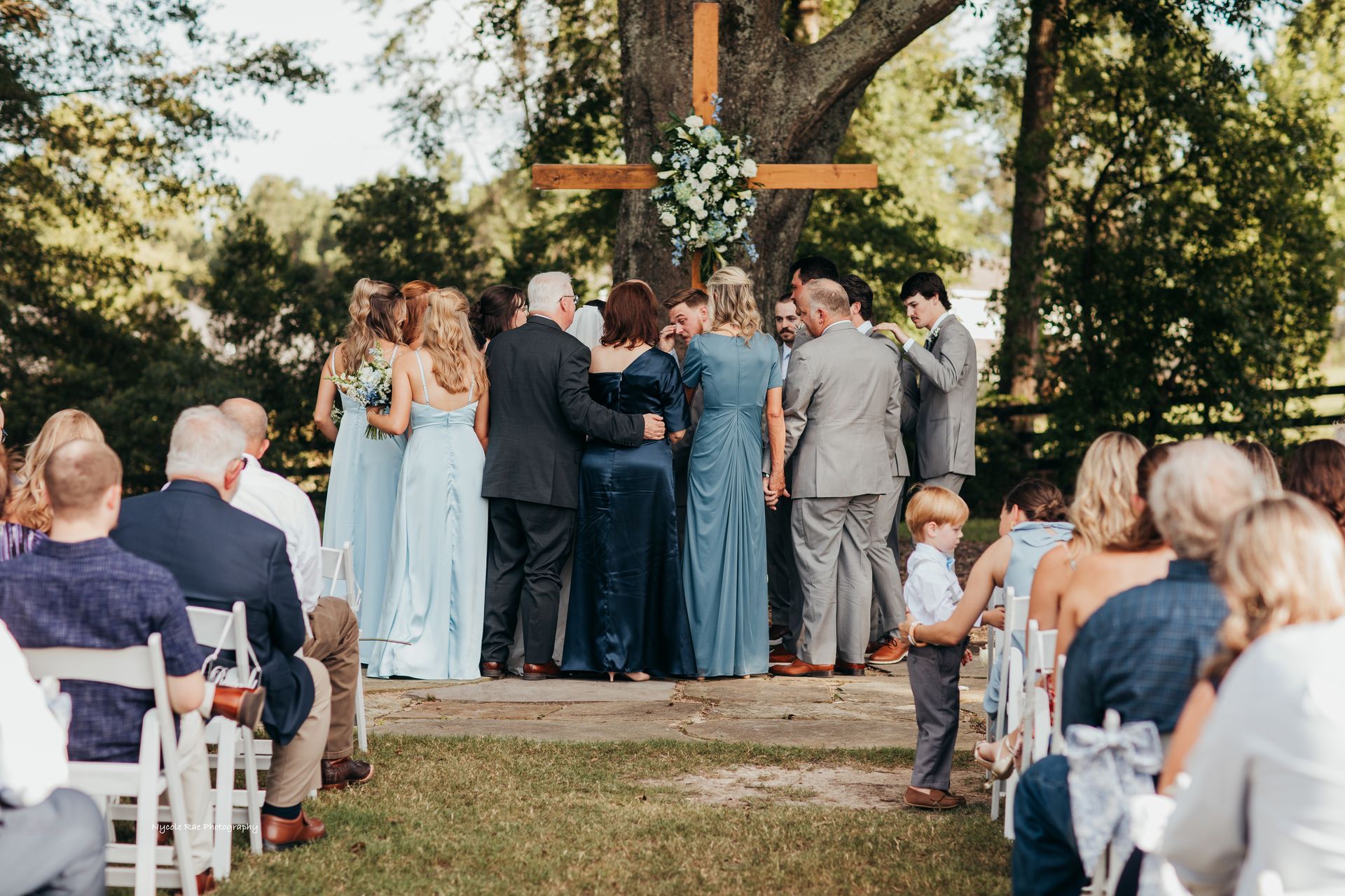 Wedding ceremony under a tree with a wooden cross, guests in attendance, couple in the center with family, blue dresses.