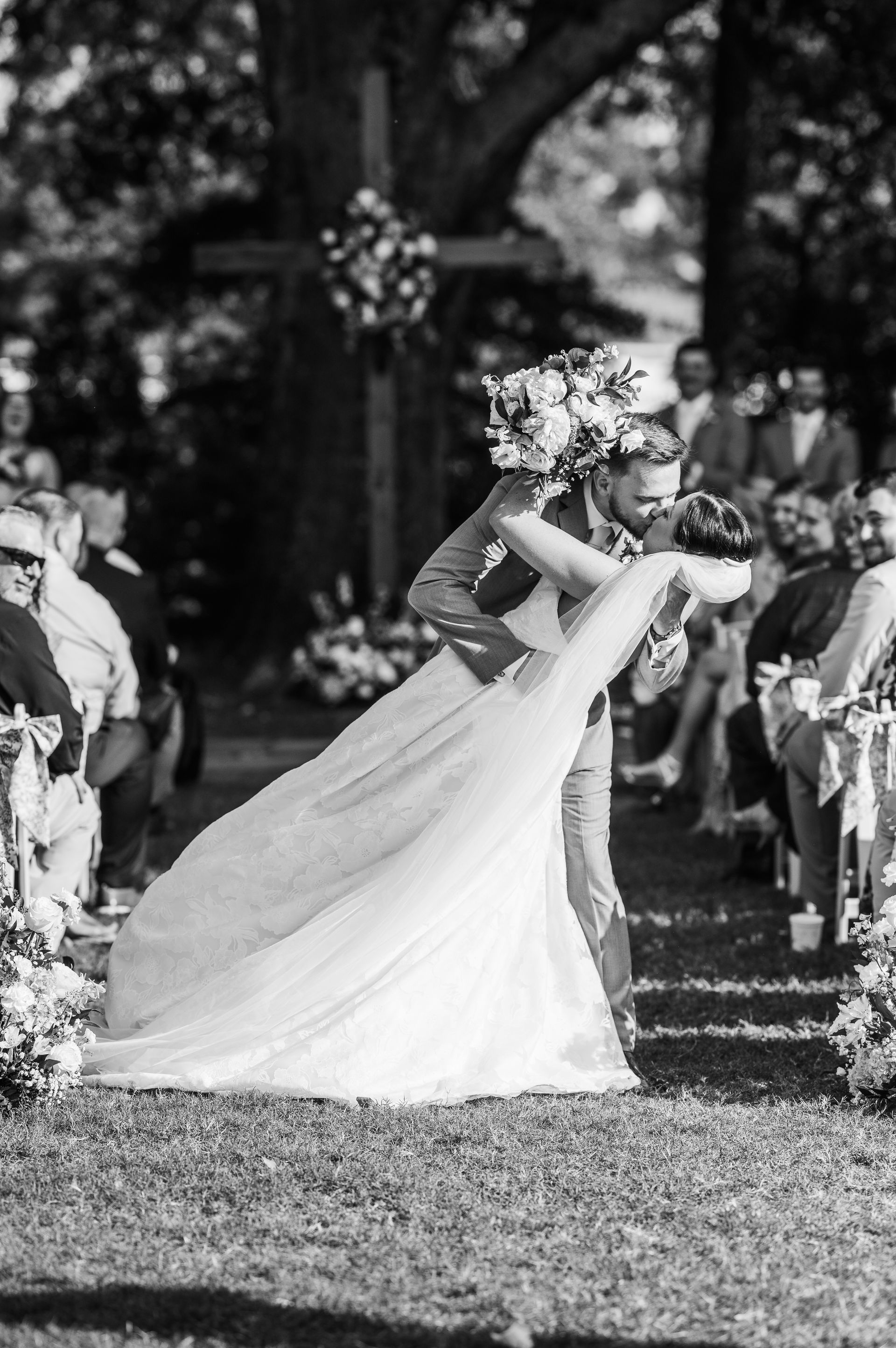 Newlyweds embrace during an outdoor wedding ceremony; bride in a flowing gown, groom in a suit, kissing.