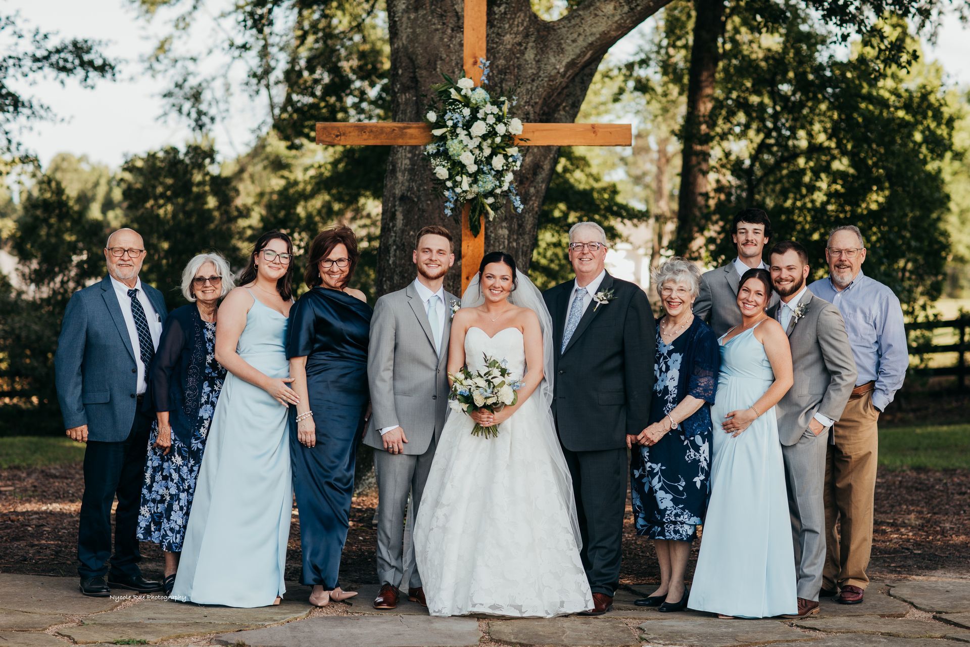 Wedding group poses in front of wooden cross. Bride and groom centered; family members on either side.