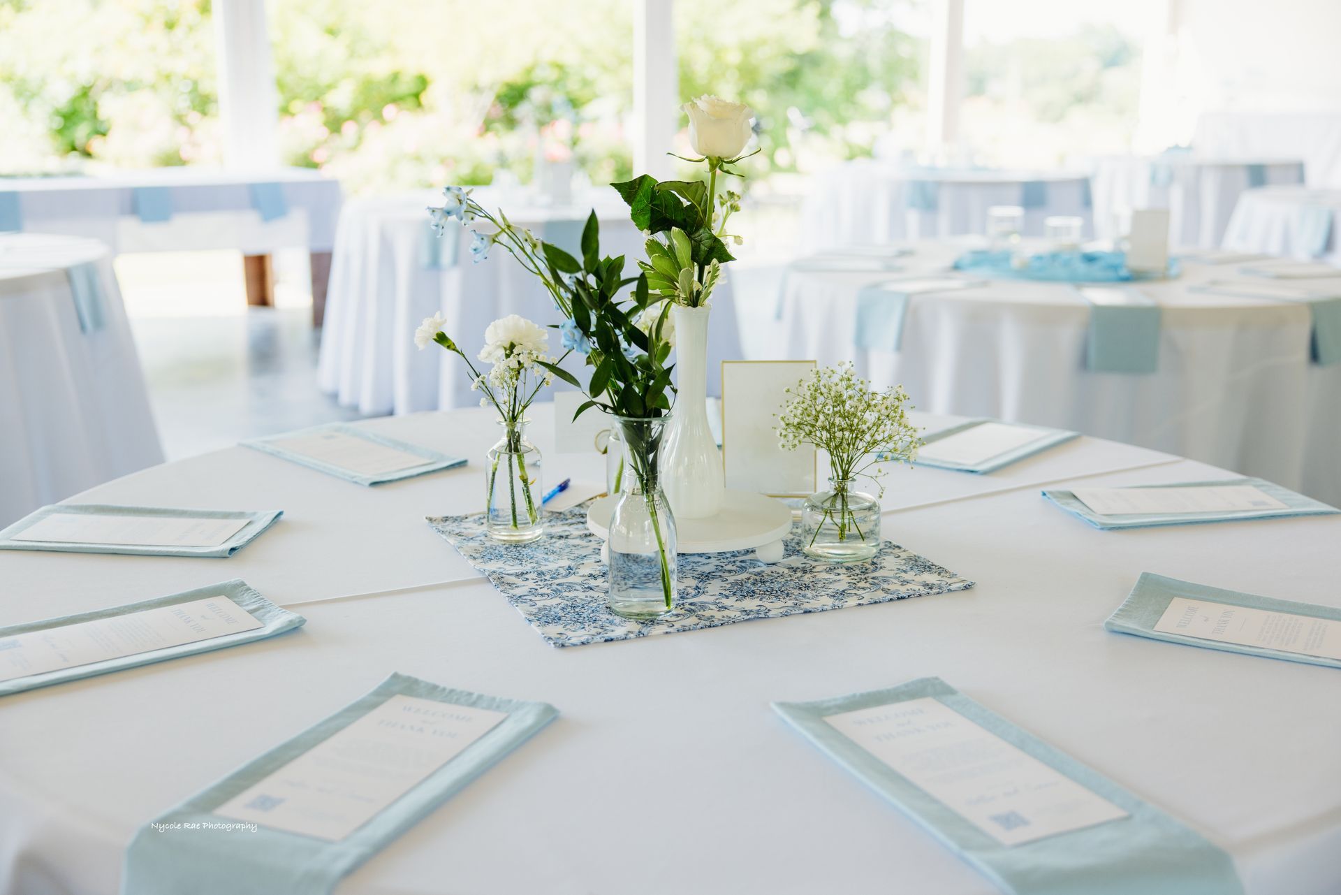 Round tables set for an event with white tablecloths, floral centerpieces, and light blue accents.