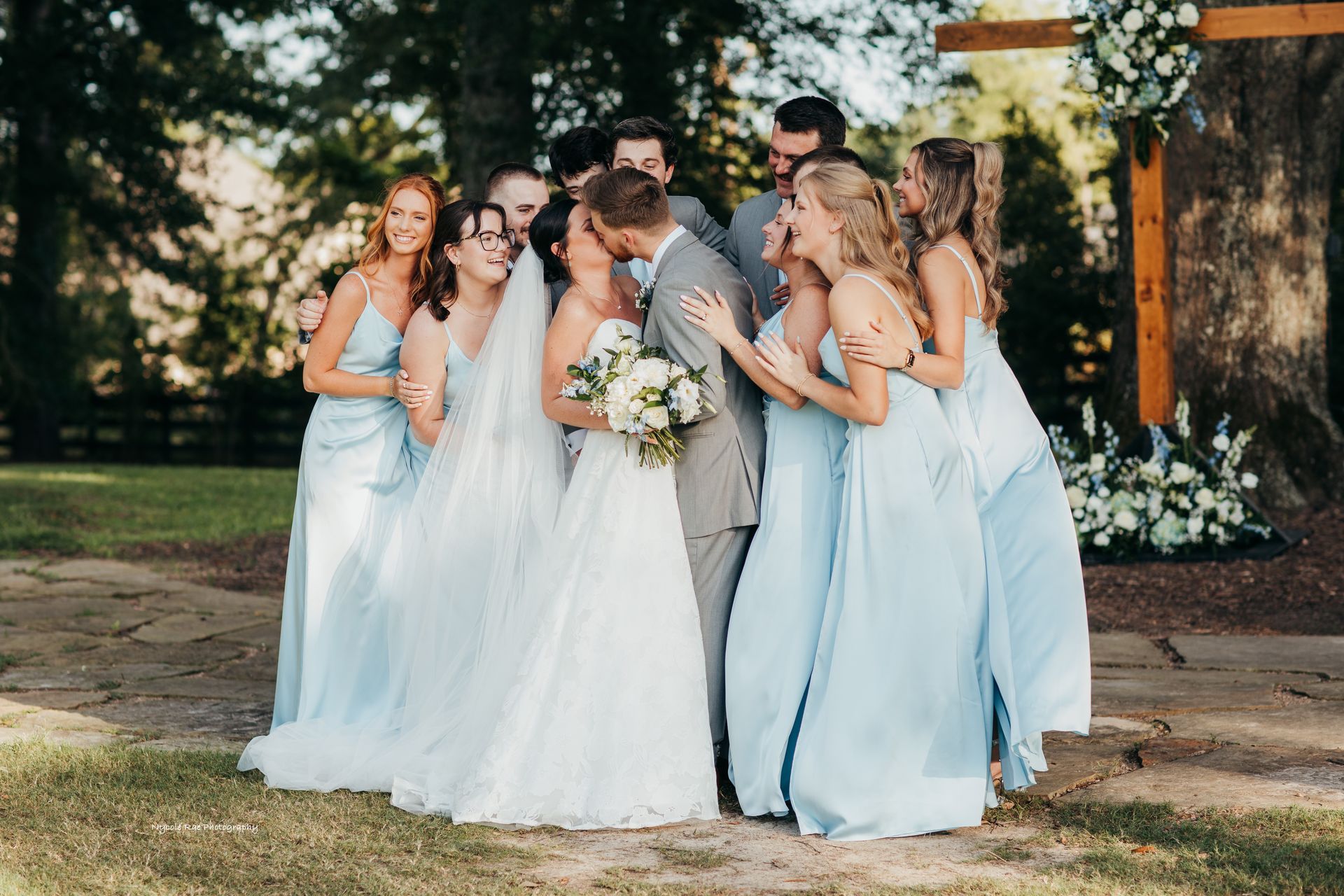 Wedding party of bridesmaids in blue dresses and groomsmen, kissing couple.