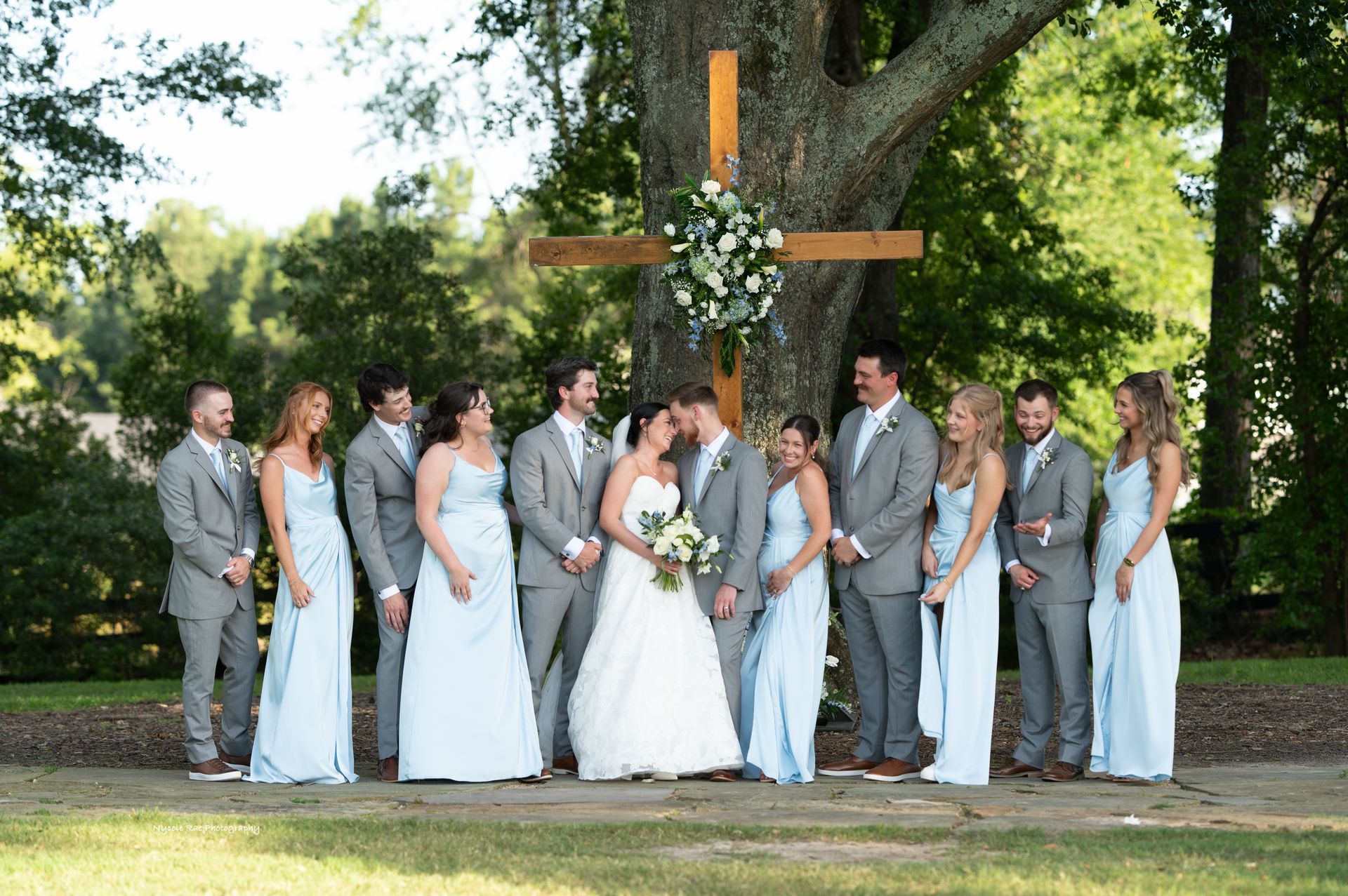 Wedding party poses in front of a tree with a wooden cross, bride and groom kiss, bridesmaids in light blue.
