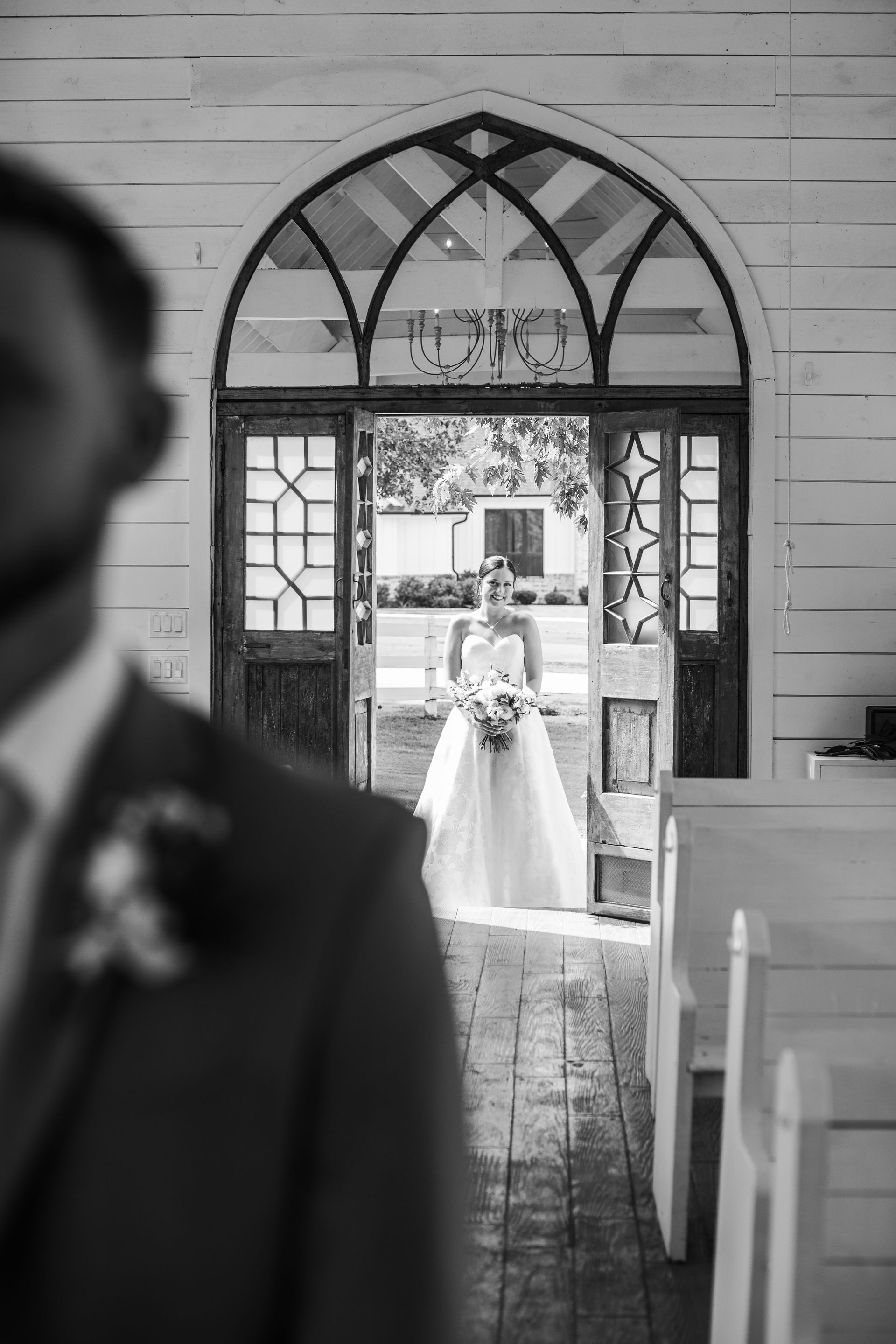 Bride in a wedding dress stands in a doorway holding a bouquet, groom in a suit is out of focus.