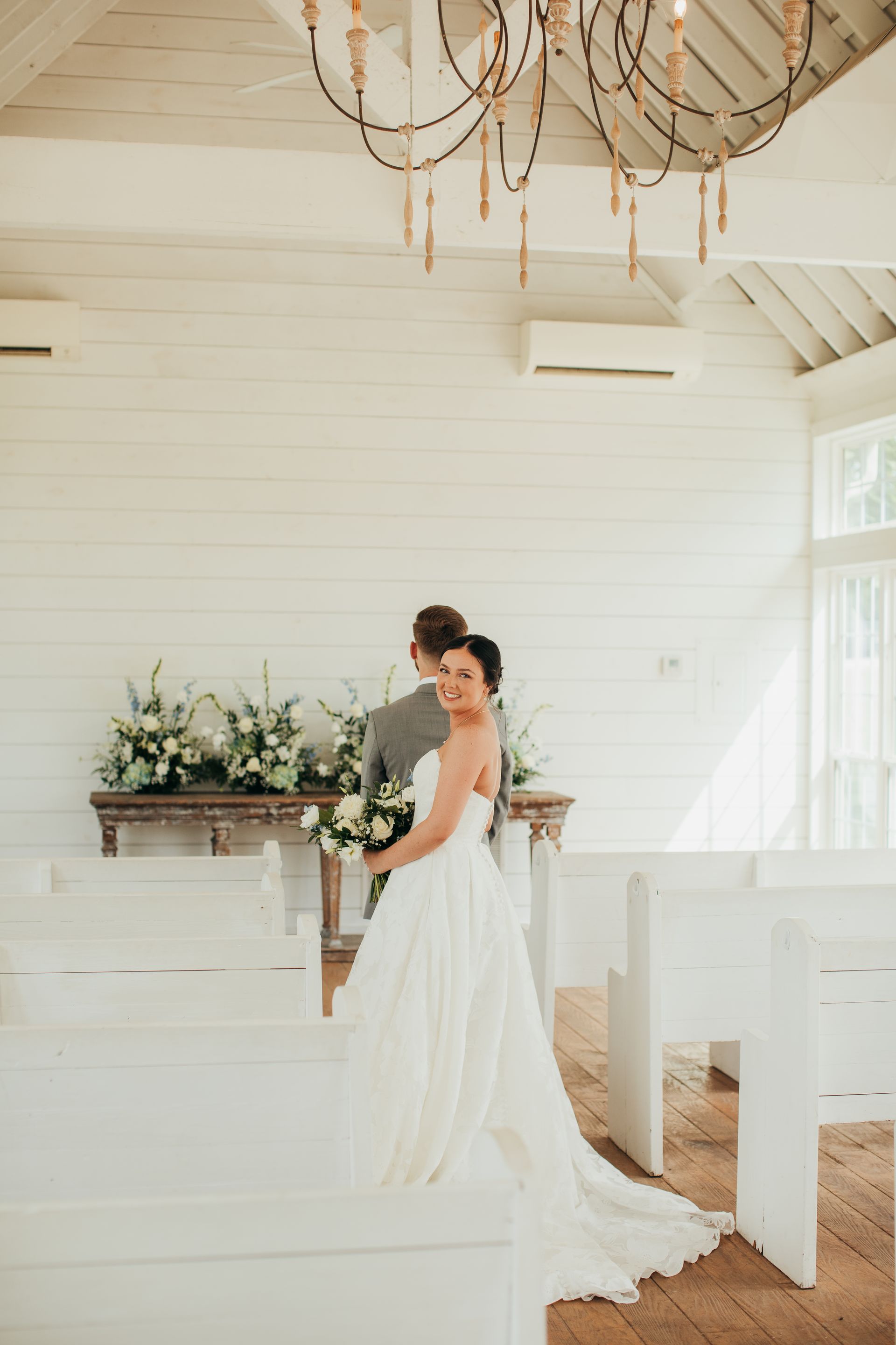 Bride and groom in a white room, she holds a bouquet, looks at the camera, he faces away.