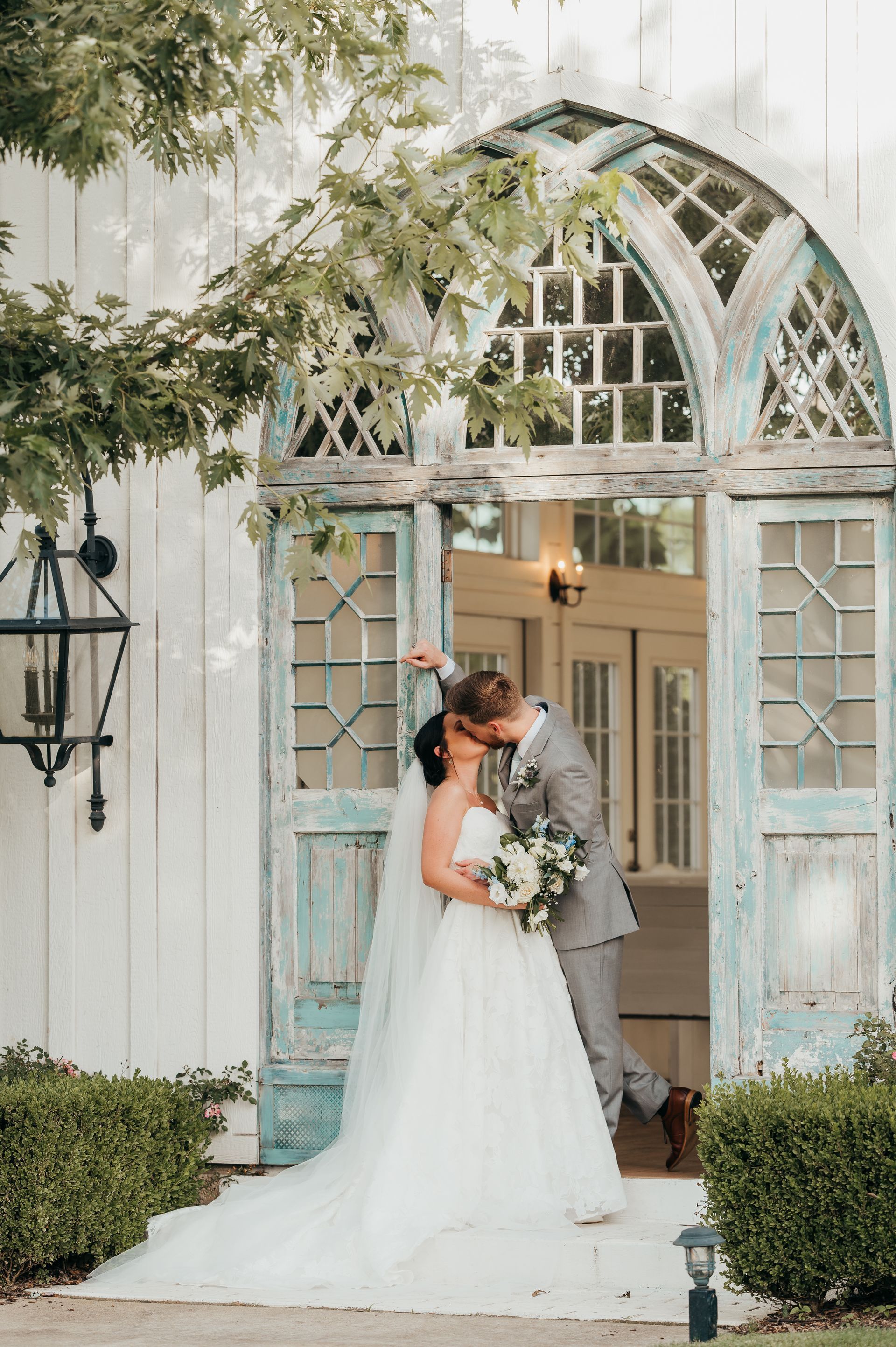 Newlyweds kissing in front of a white door with blue trim; woman in a white gown, man in a grey suit.