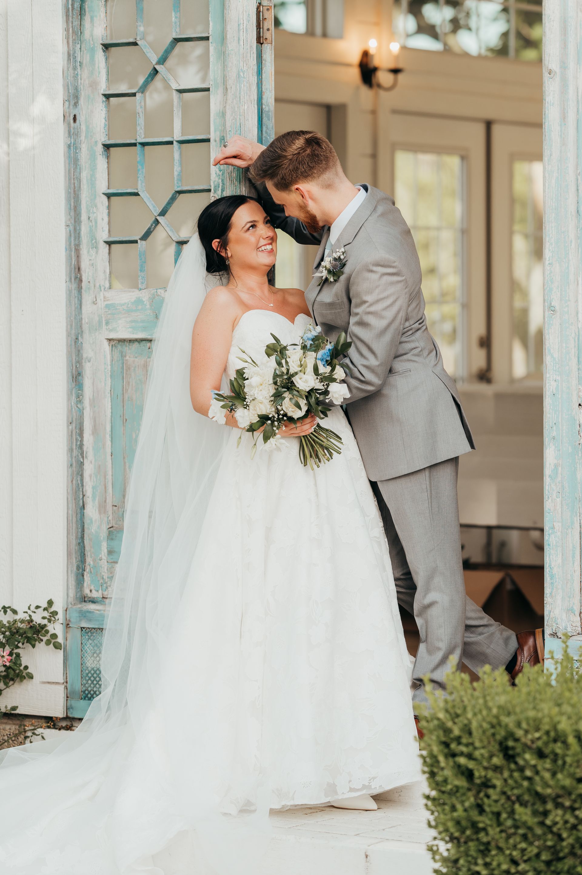 Bride and groom embracing, smiling, standing in doorway. Bride in white gown, veil, holding bouquet. Groom in gray suit.
