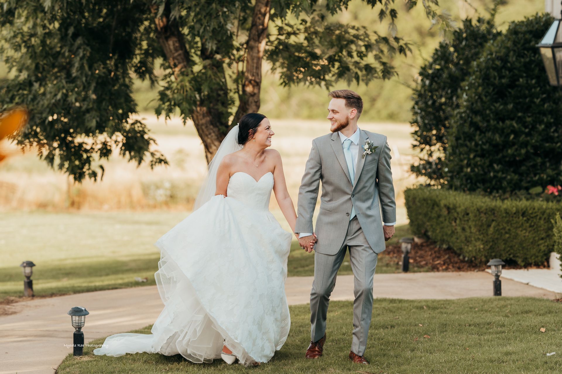 Bride and groom walking, holding hands, smiling. She wears a white gown, he wears a gray suit, outdoors.