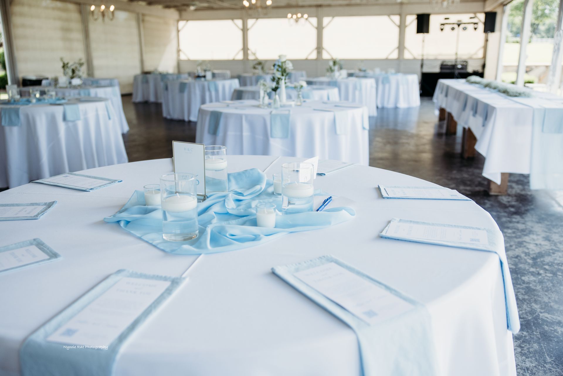 Wedding reception tables set with white tablecloths, blue runners, and centerpieces, in a light-filled venue.