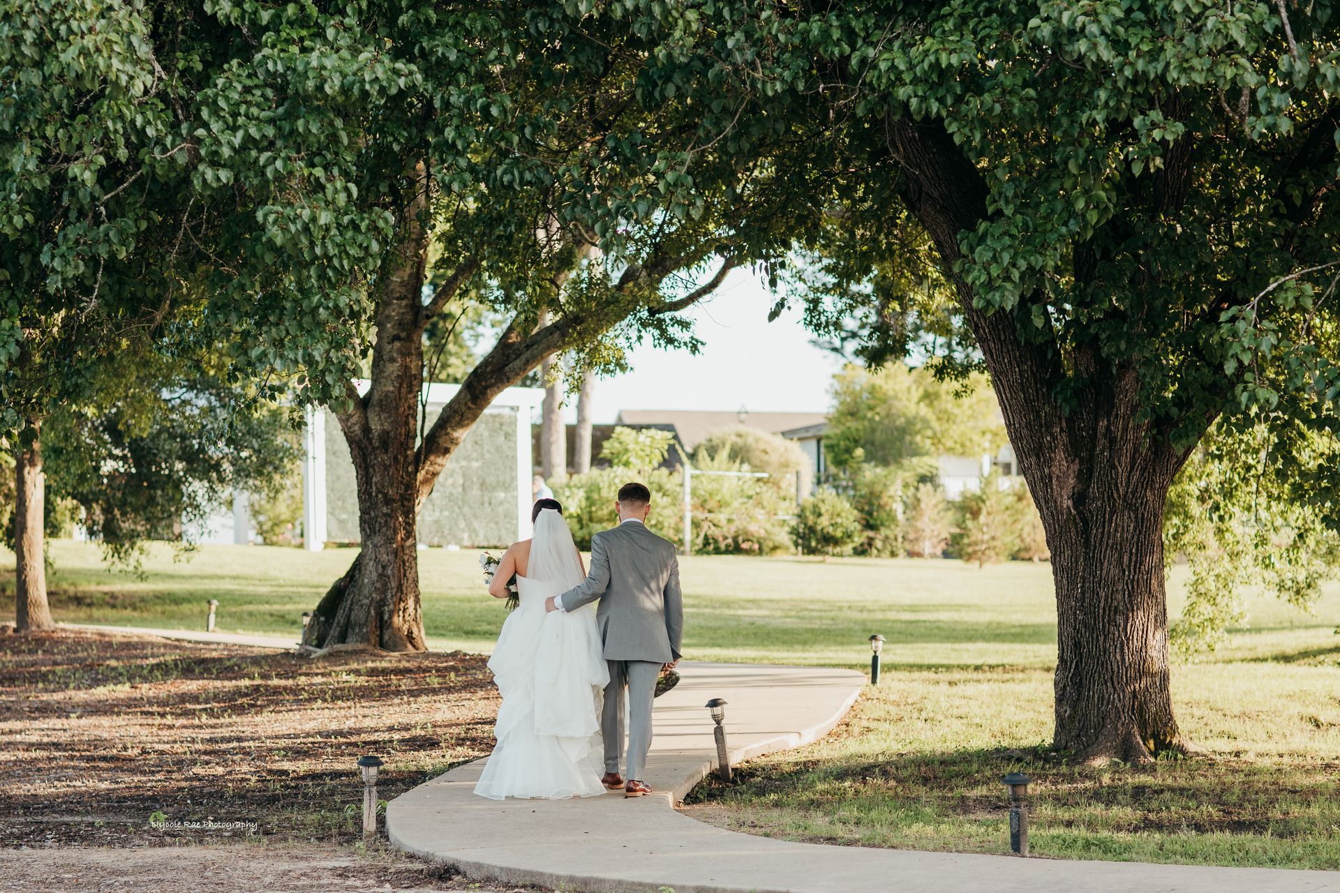 Bride and groom walk away from the camera on a path under trees, towards a building with a white exterior.