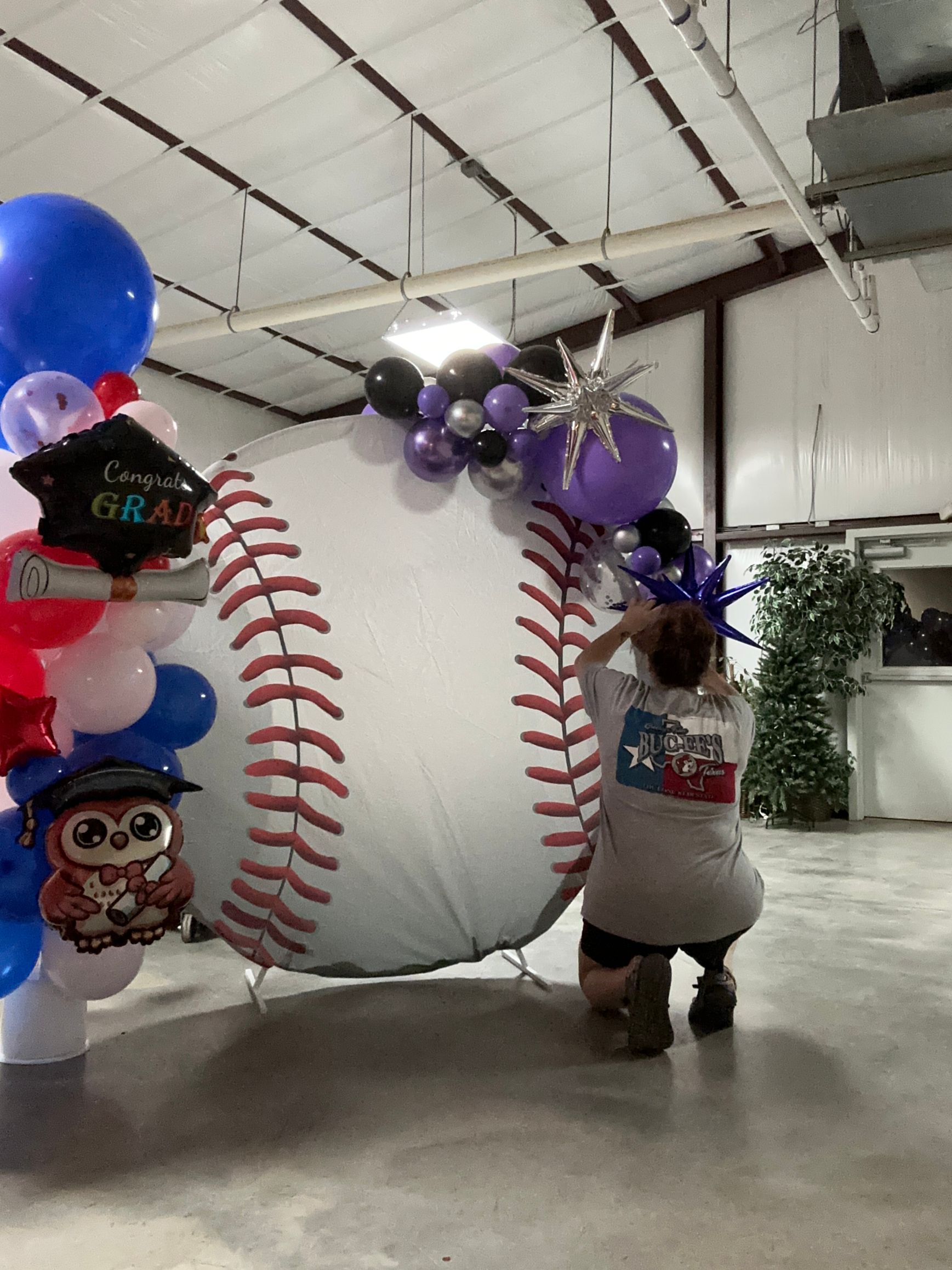 Person adjusting a large baseball-themed balloon arch with graduation balloons in a gymnasium.