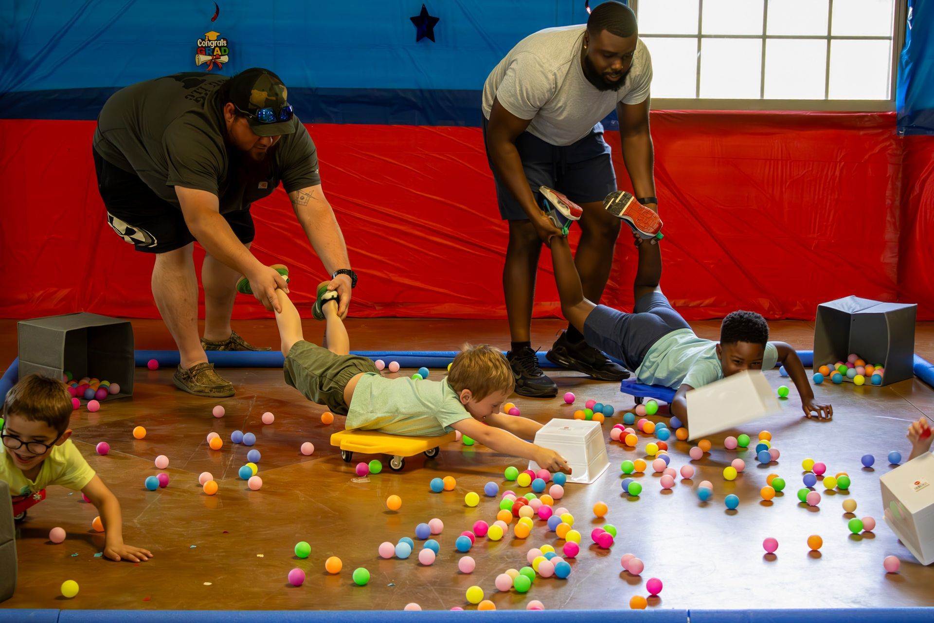 Children play a game with adults; they lie on rolling boards to gather colorful balls on a floor.
