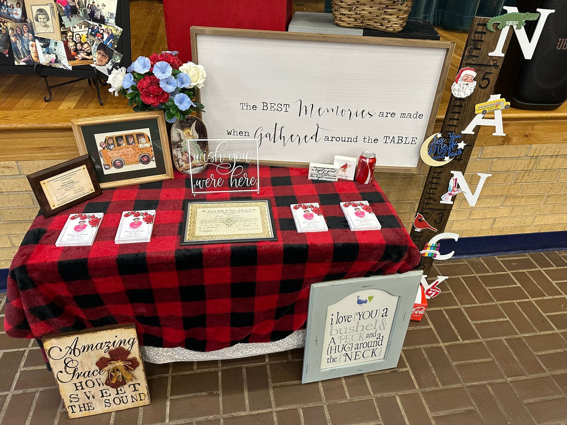 A craft fair booth with red and black checkered tablecloth, signs, and floral arrangements.