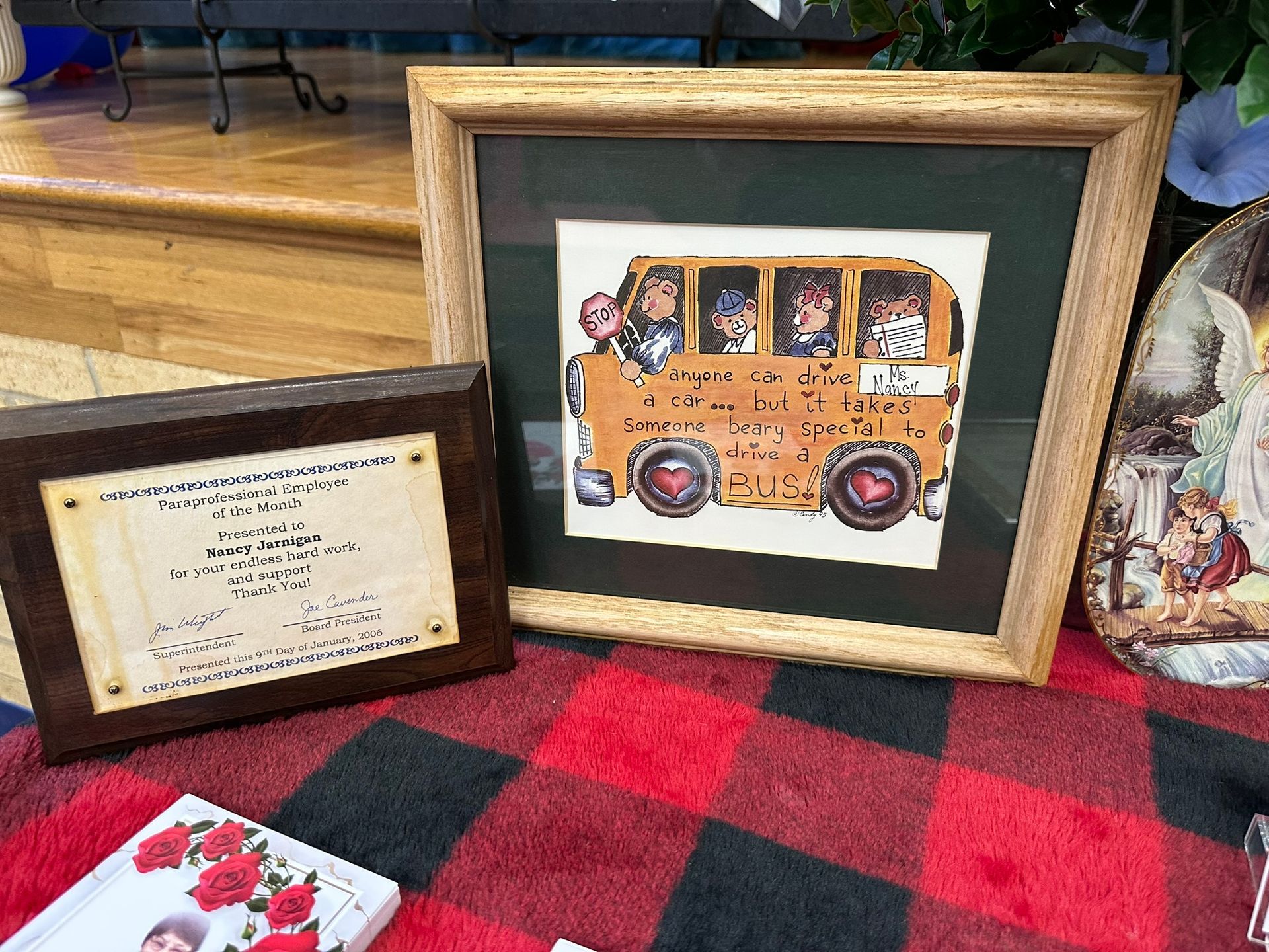 Framed artwork of a school bus with children. Next to it, a framed document. Both sit on plaid fabric.