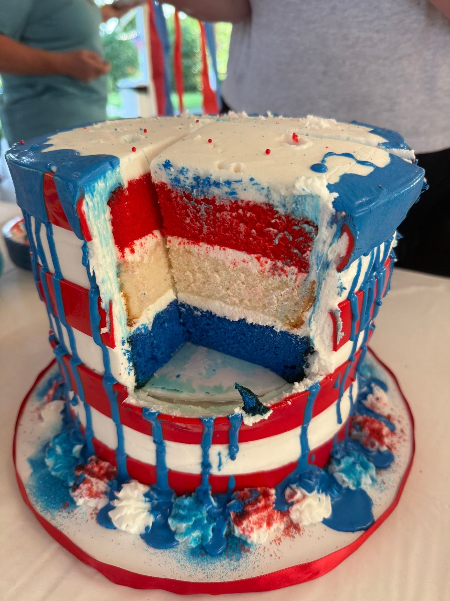 Patriotic top hat cake: red, white, and blue layers, cut open, with dripping frosting and decorations.