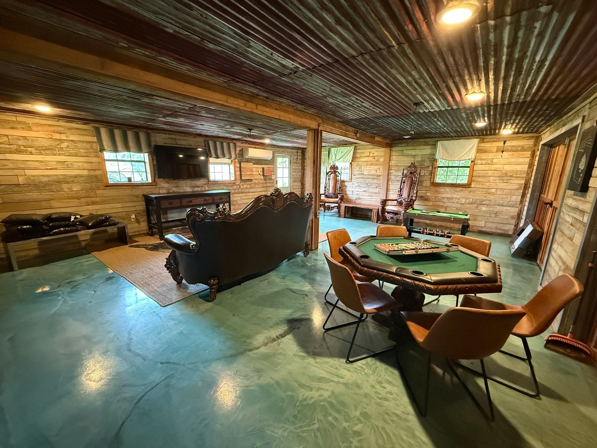 A rustic game room with a poker table, sofa, and wood-paneled walls. The floor is a teal epoxy.