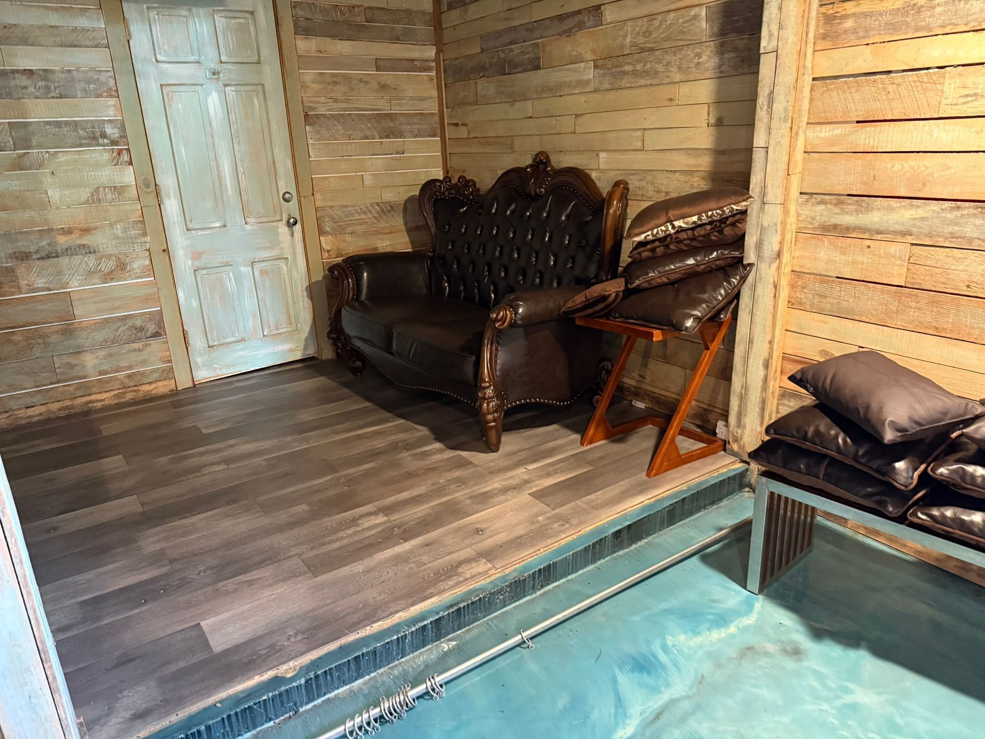 A rustic room with wood-paneled walls, leather couch, recliner, door, and a partially visible pool.