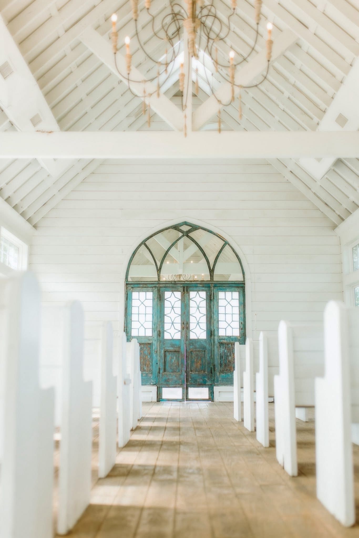 White chapel interior with wooden pews, teal doors, and a chandelier.
