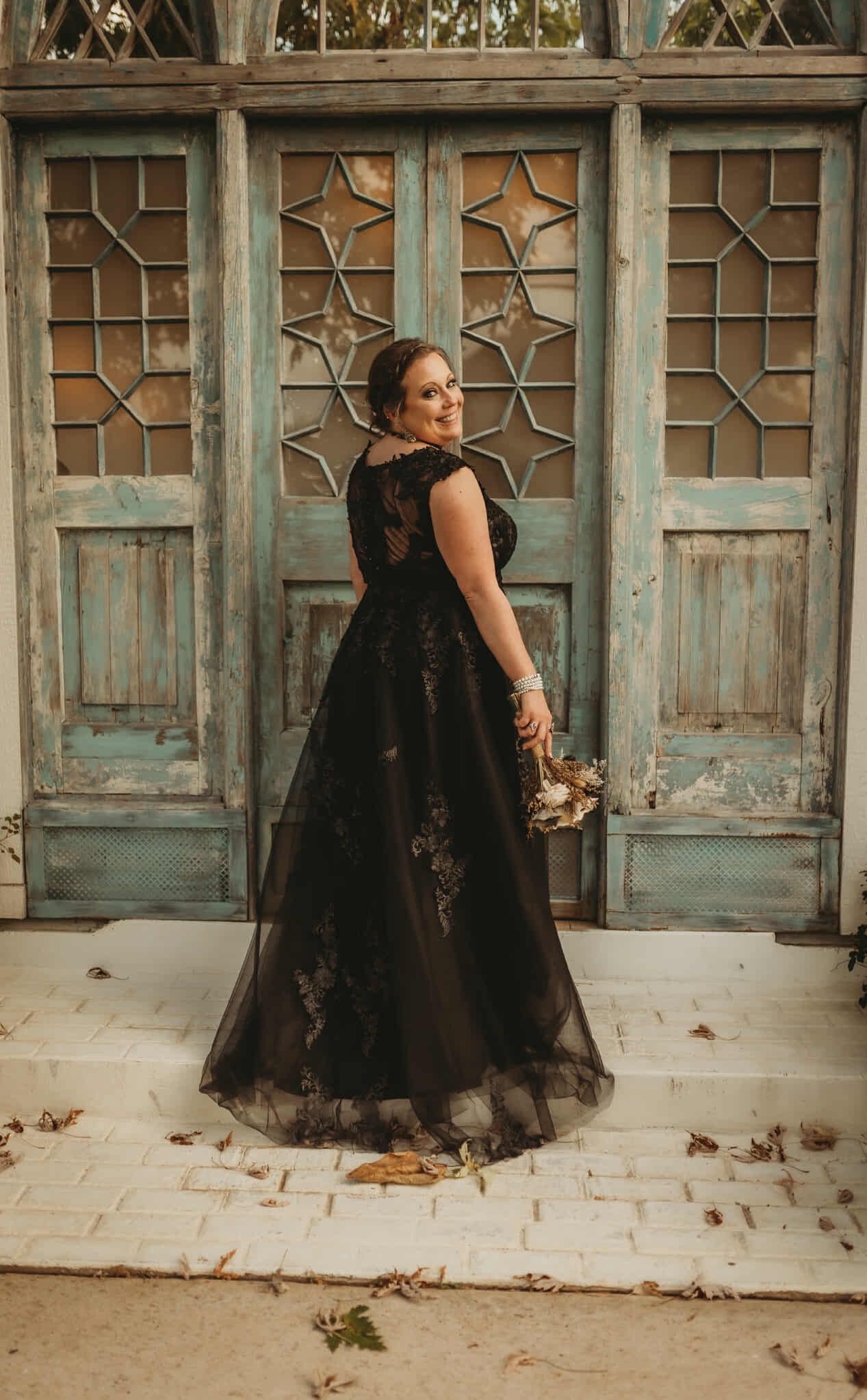 Woman in black dress, holding bouquet, poses in front of weathered blue doors, smiling over shoulder.