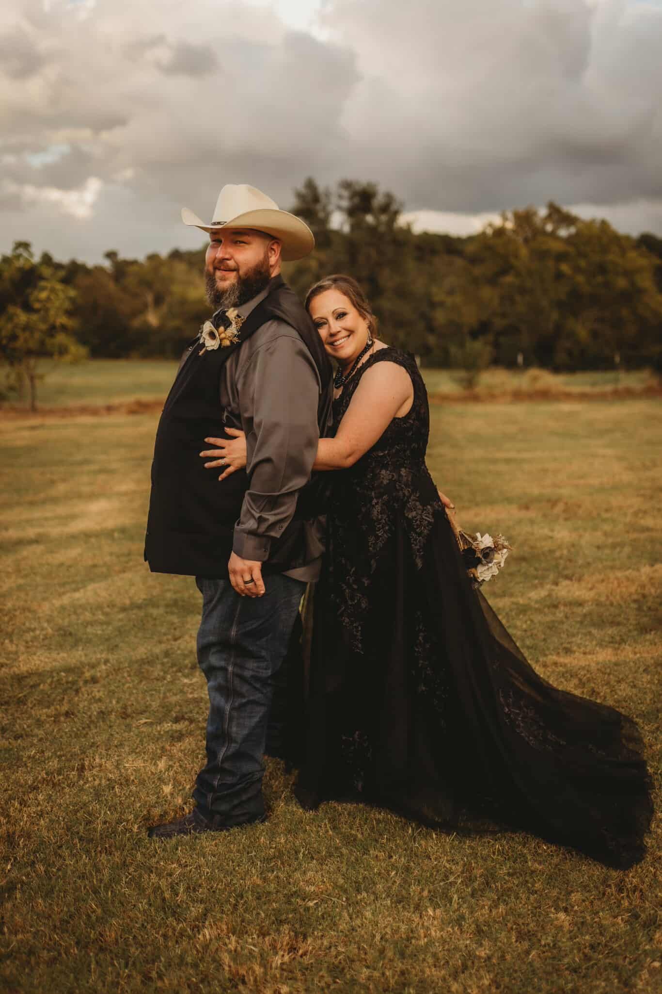 Couple in wedding attire embrace outdoors. Woman in black dress, man in cowboy hat and vest.