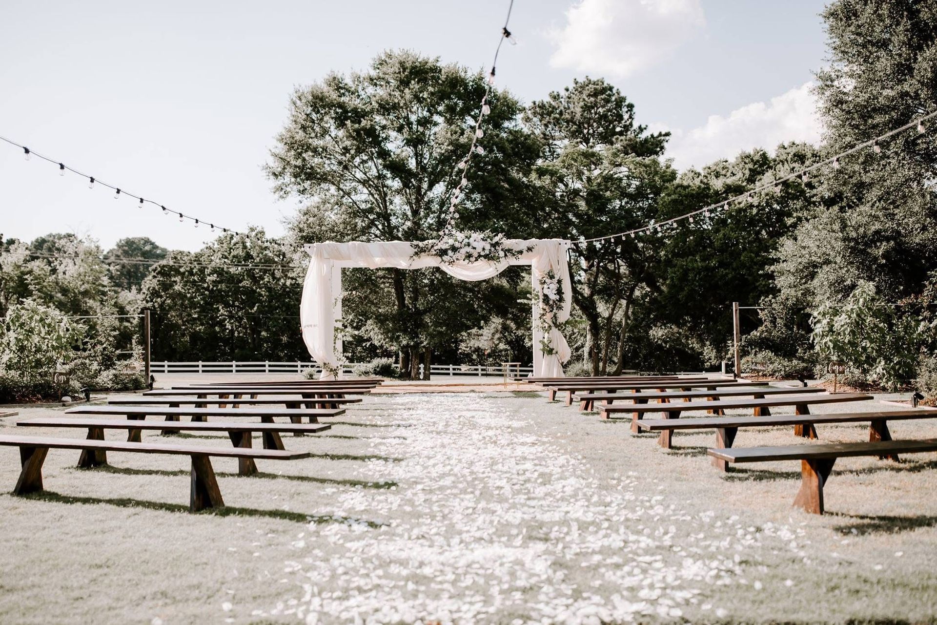 Outdoor wedding ceremony setup with white floral arch, benches, and petal-strewn aisle.