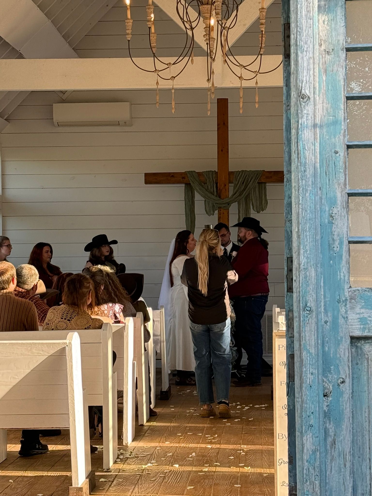 Wedding ceremony in a rustic white chapel with a cross, people are seated and standing; a photographer is present.