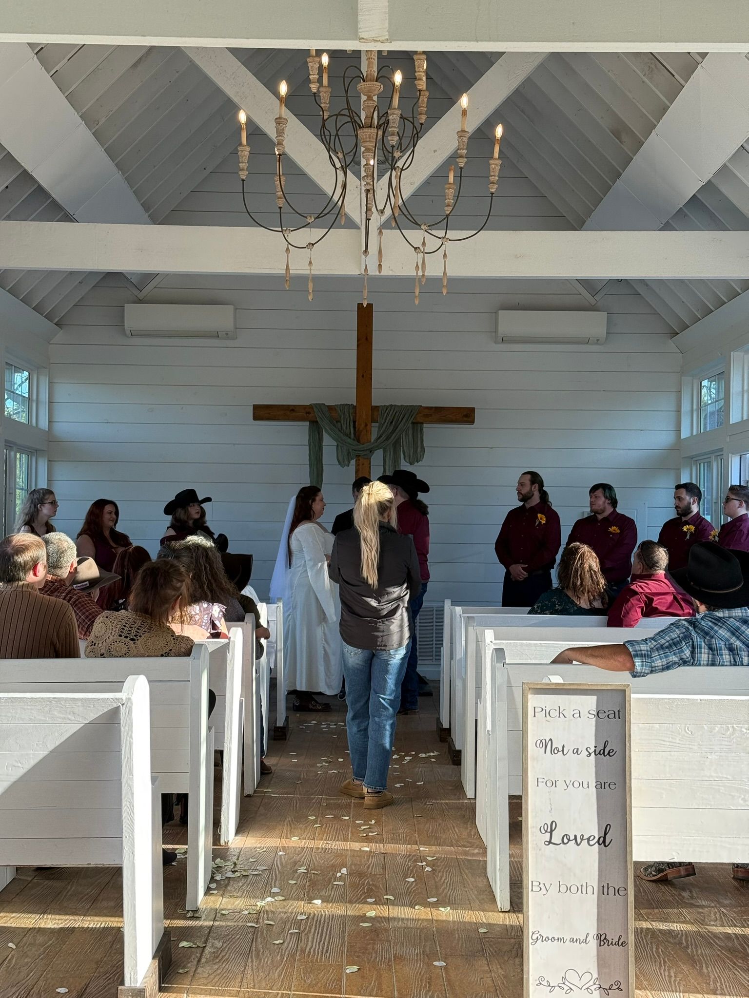 Wedding ceremony in a white chapel. People are gathered near the altar with a cross, a chandelier hangs above.