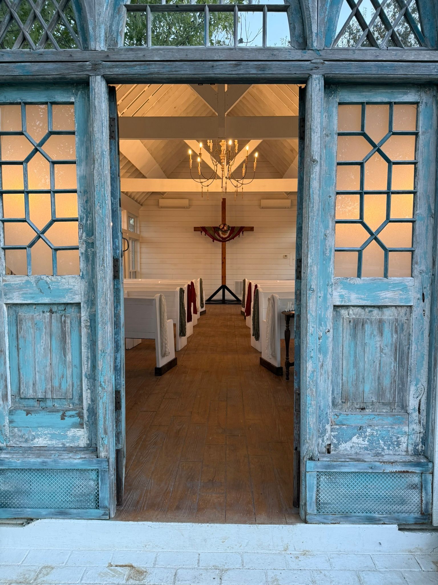 Open, distressed blue doors frame a bright chapel interior with a crucifix and rows of seating.