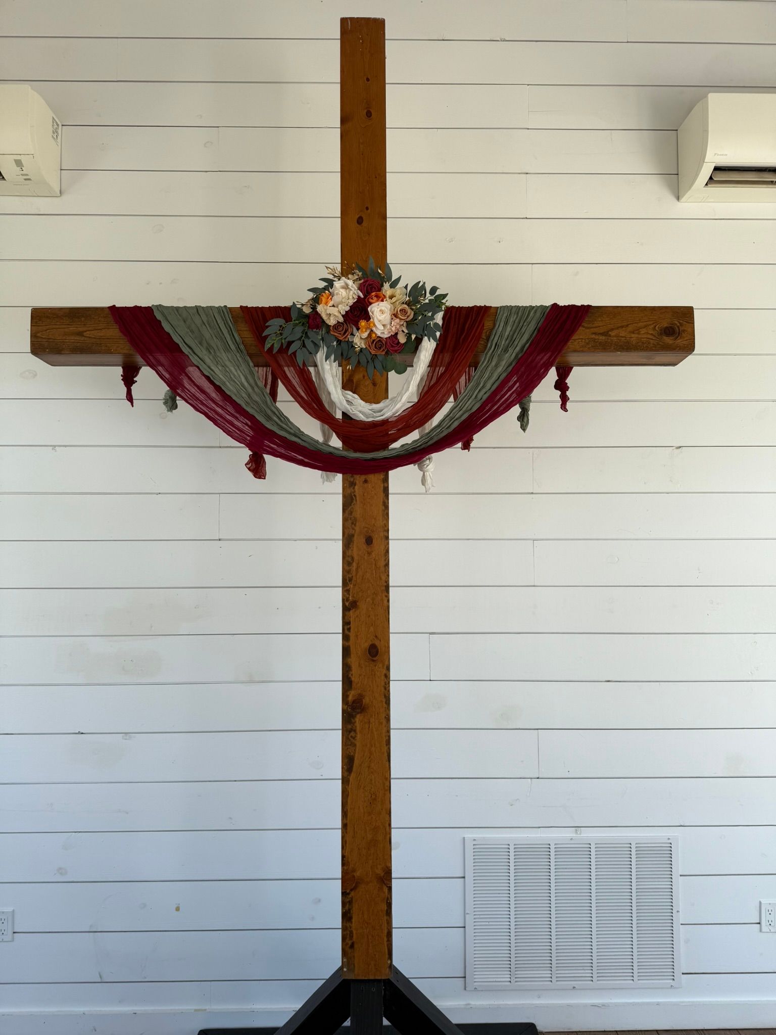 Wooden cross decorated with flowers and a burgundy and green draped fabric against a white wall.
